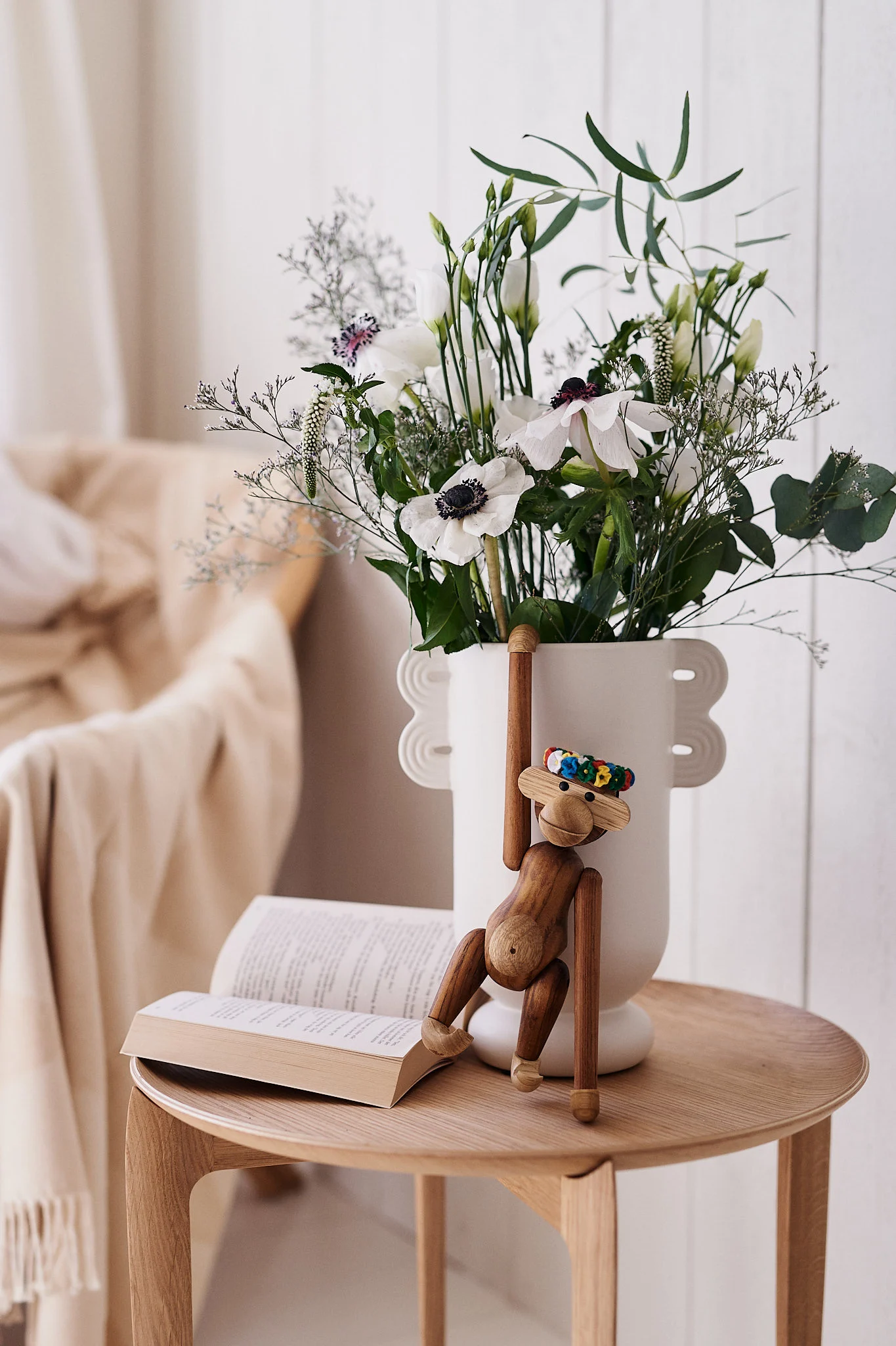 A wooden monkey toy hangs from a white vase filled with white flowers next to an open book on a wooden table.