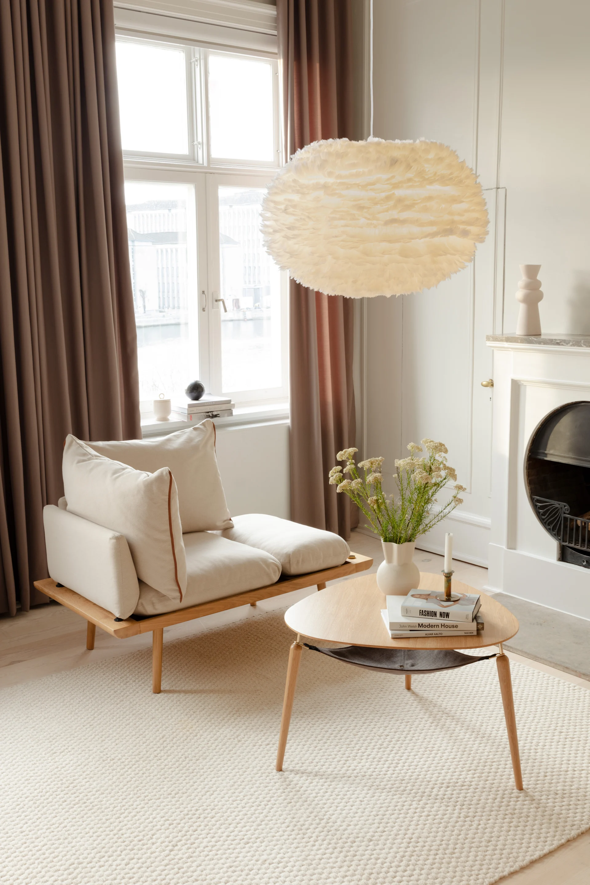 A bright living room with a cream sofa, wooden coffee table, flowers, and a large feather pendant lamp.