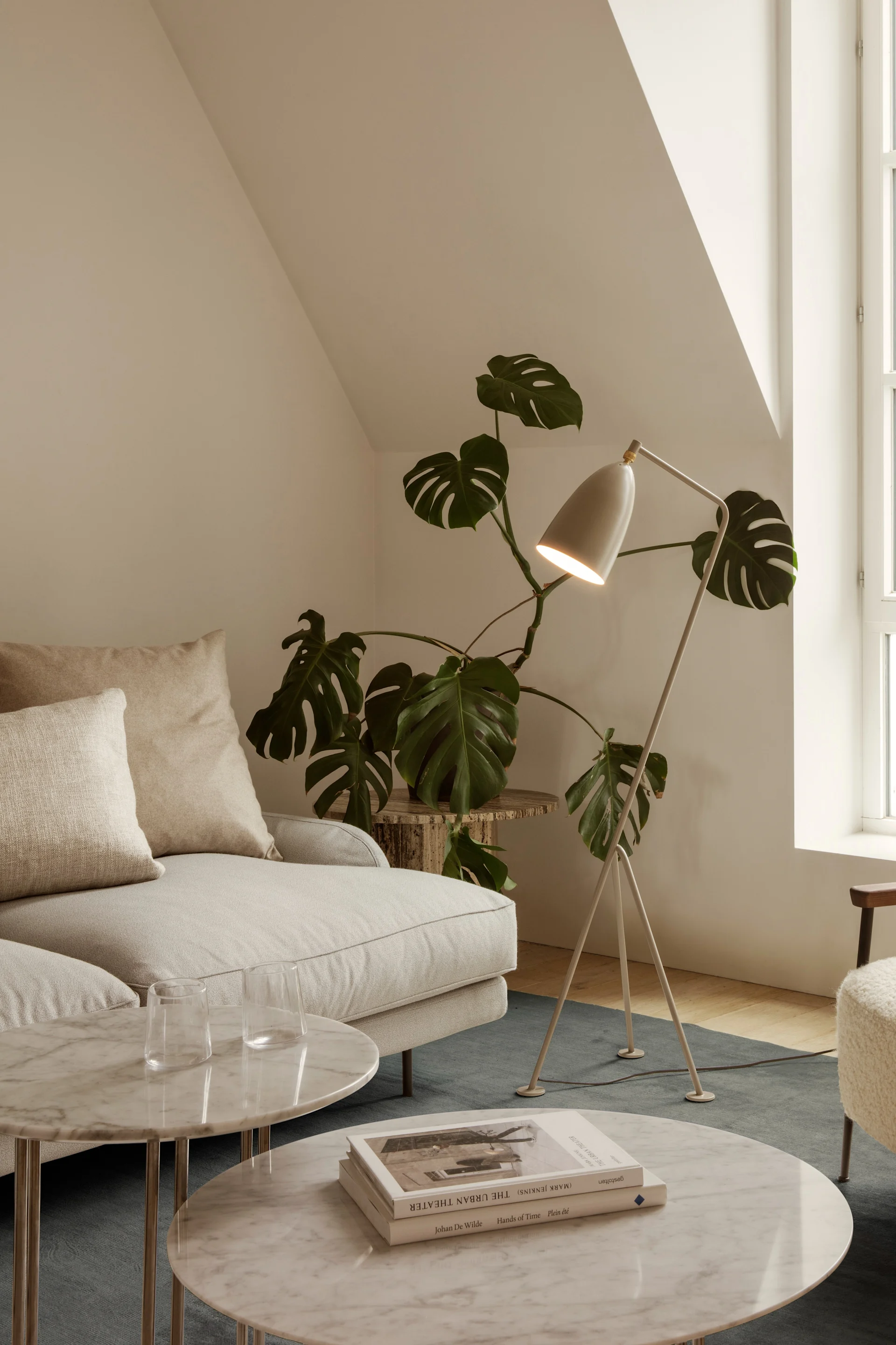A bright living room with a cream sofa, marble coffee tables, a floor lamp, a plant, and a fluffy armchair by an arched window.