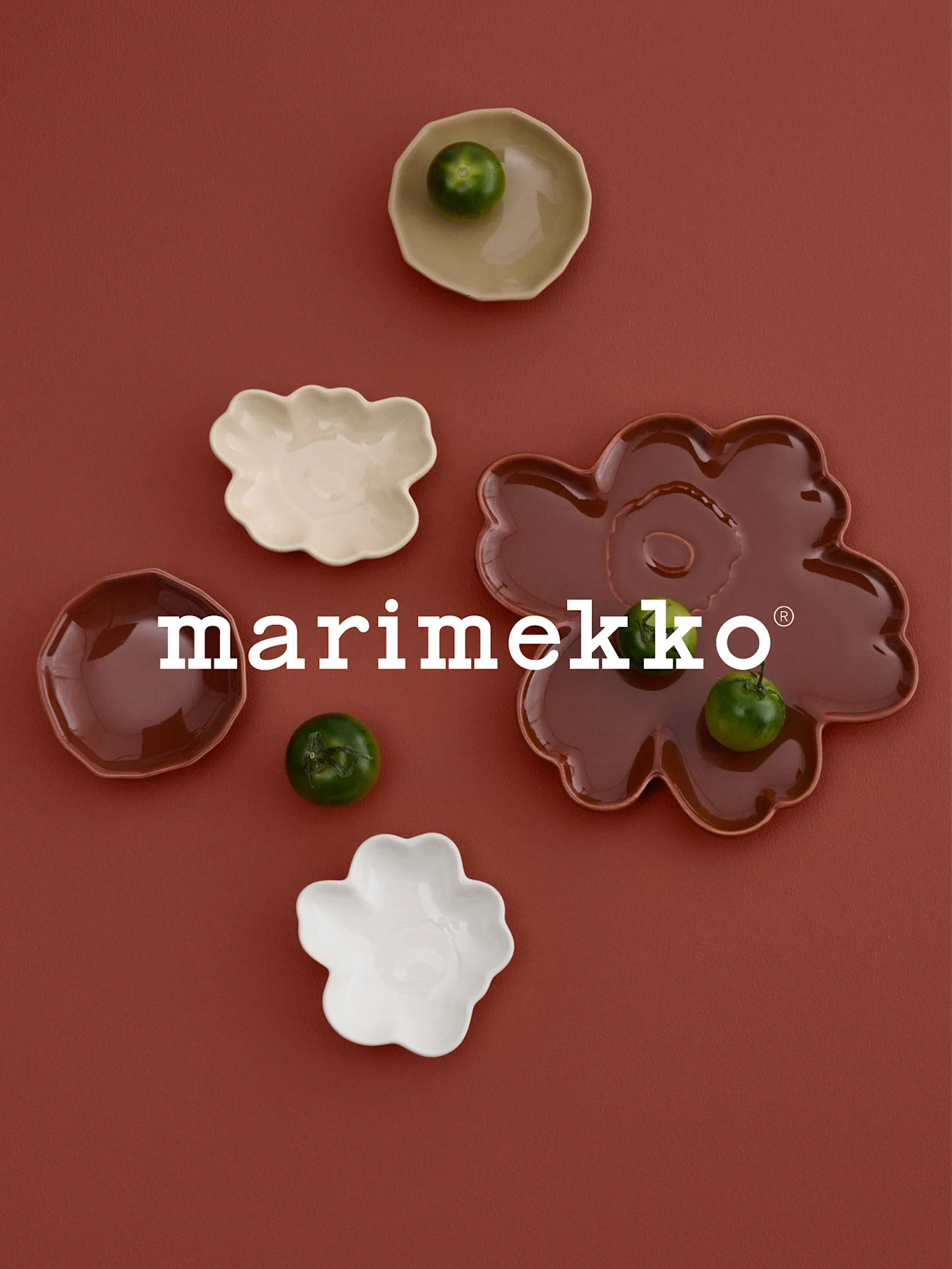 White 'marimekko' logo, flower-shaped dishes (white, cream, reddish-brown) with green fruits, on a rust surface.
