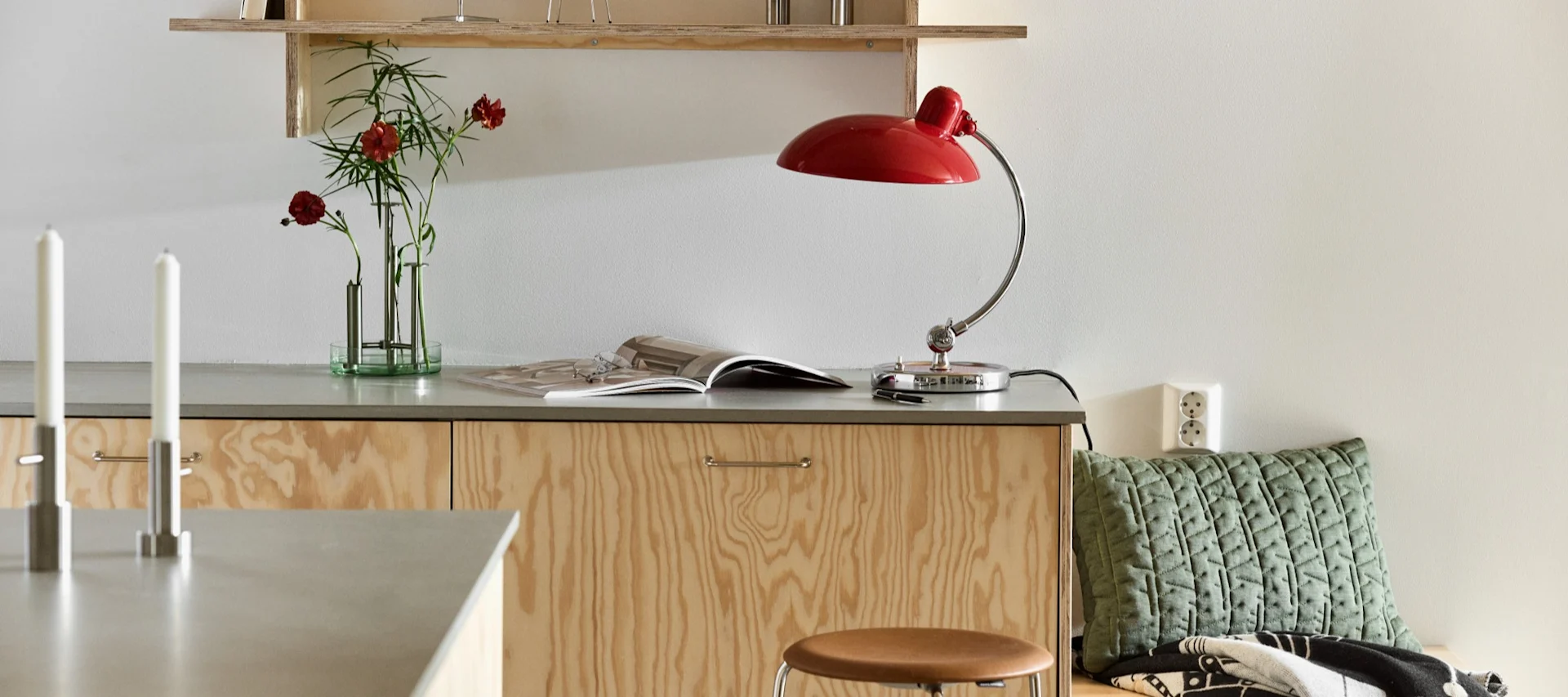 Here you see the Kaiser table lamp in ruby red with the Ikeru vase in brushed steel, both from Fritz Hansen, in a modern plywood kitchen.