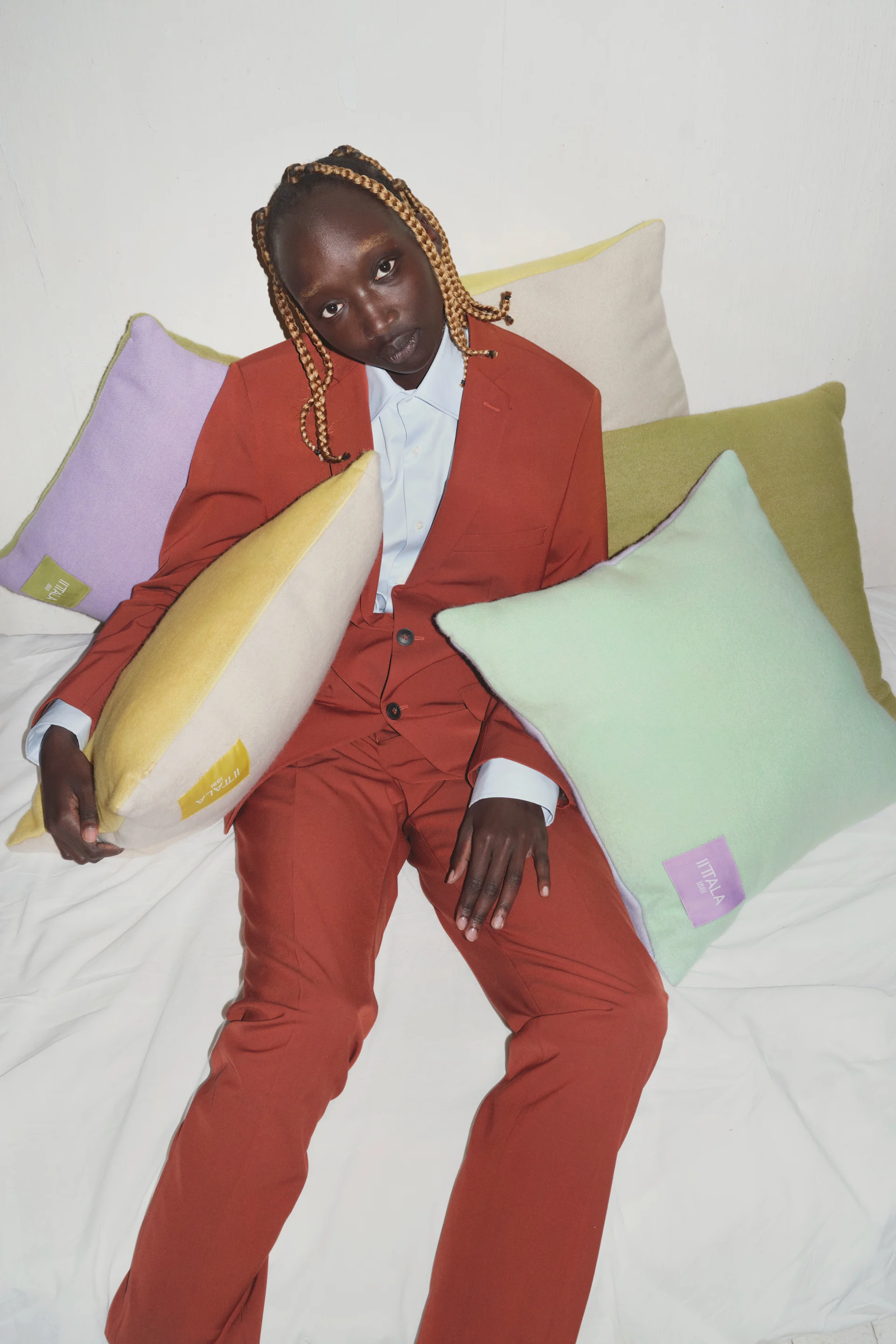 Person with light brown braids in rust-colored suit on white bed with pastel Iittala pillows.