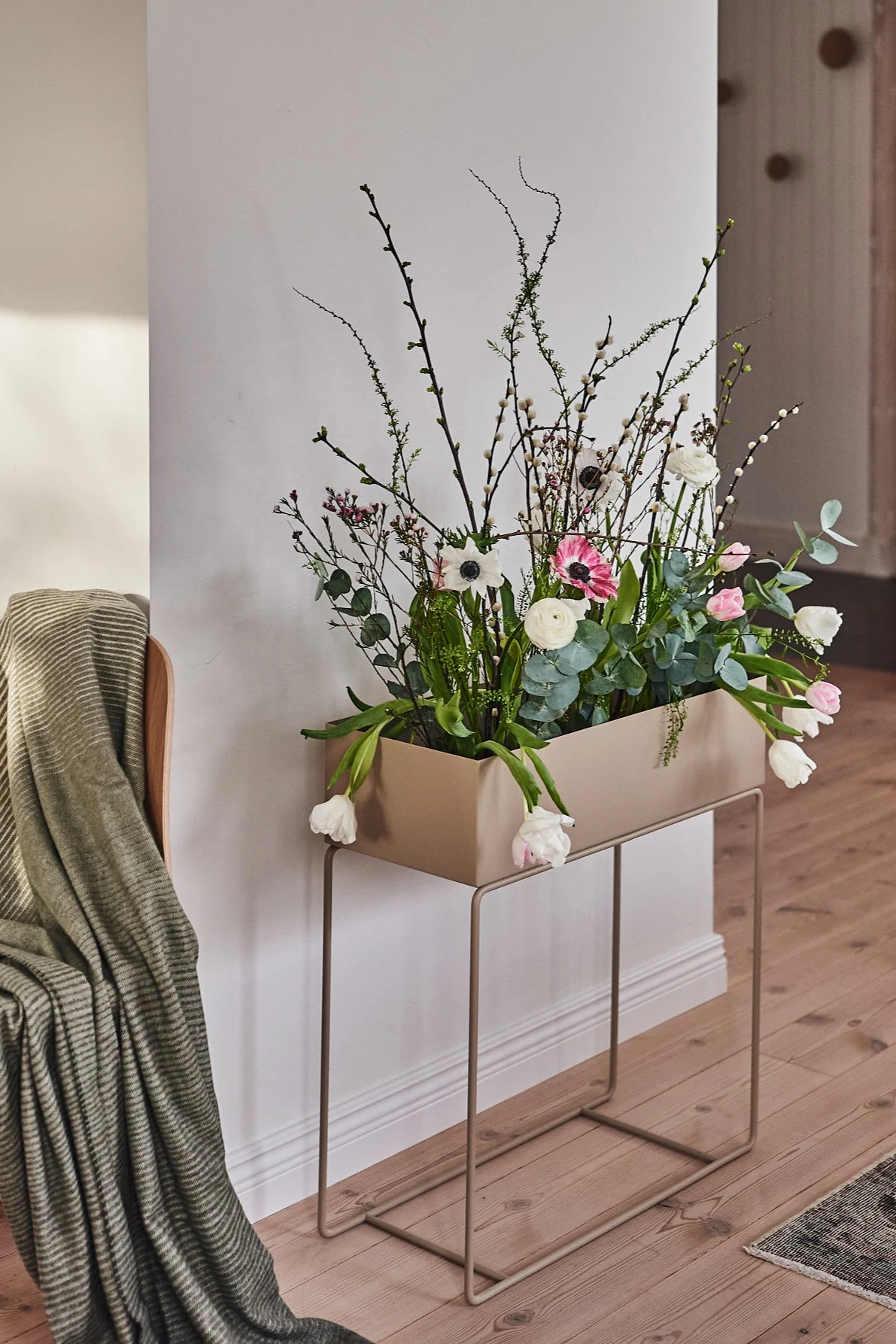 A rectangular beige planter on a minimalist stand filled with white and pink flowers, eucalyptus, and budding branches, next to a chair with a green striped blanket.