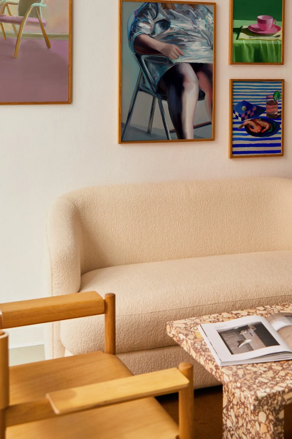 A cream bouclé sofa, wooden armchair, and terrazzo coffee table with an open magazine, with framed paintings on the wall.