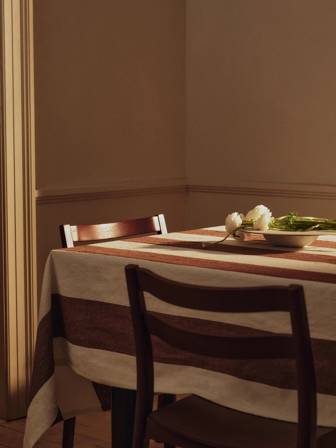Dining table with a striped tablecloth, two wooden chairs, and white flowers in a small bowl.