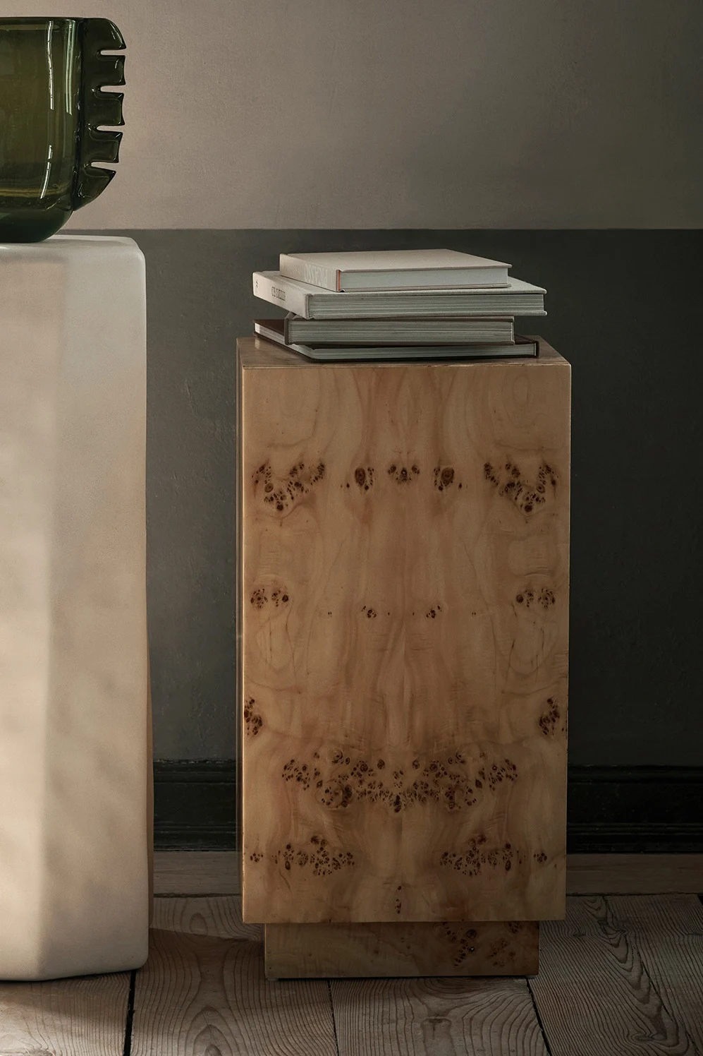 A burled wood rectangular pedestal with a stack of books, next to a light octagonal pedestal holding a dark green ribbed vase.