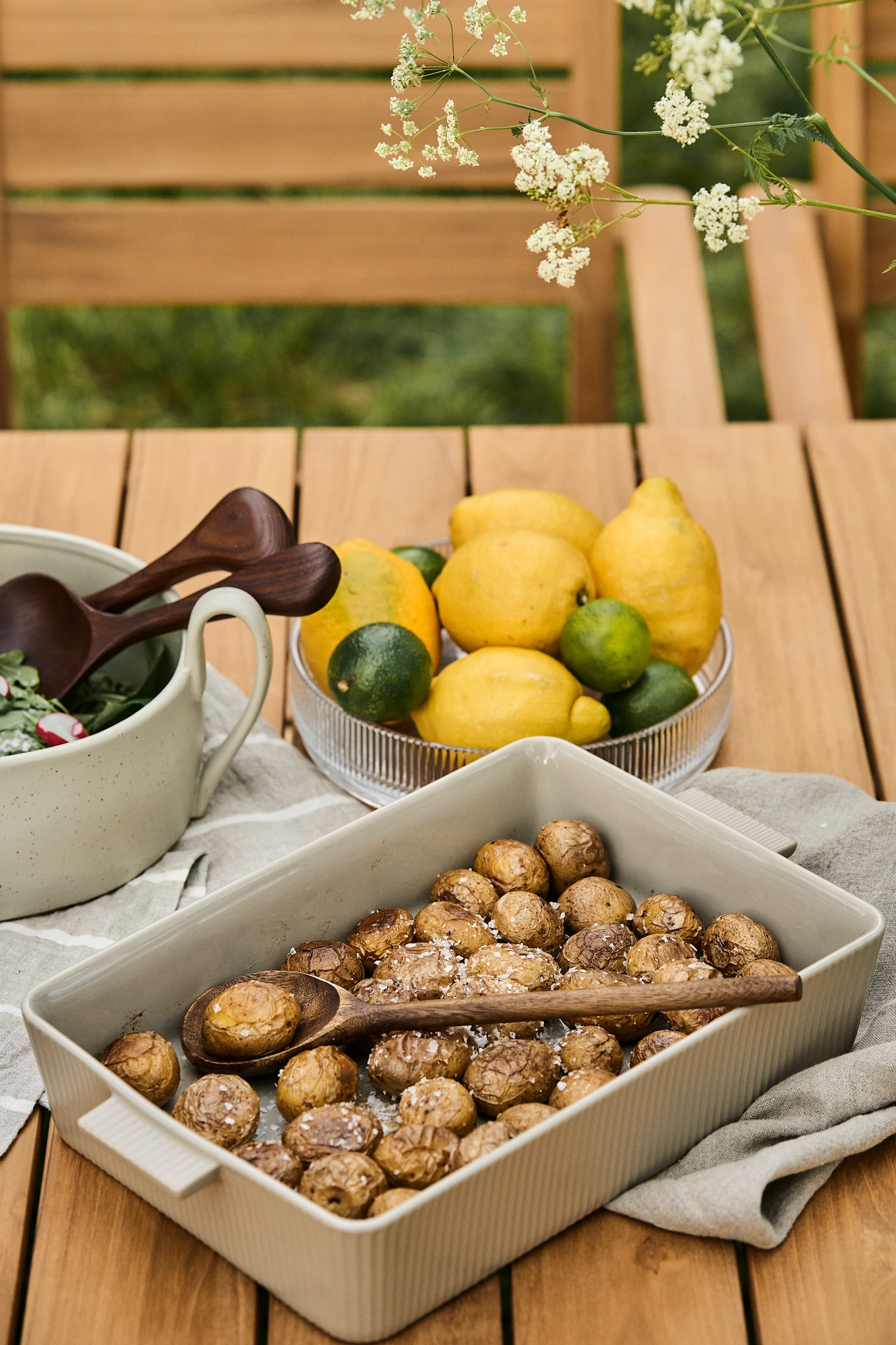 Roasted baby potatoes with salt in a baking dish, salad bowl with wooden spoons, and a bowl of lemons and limes on an outdoor wooden table.