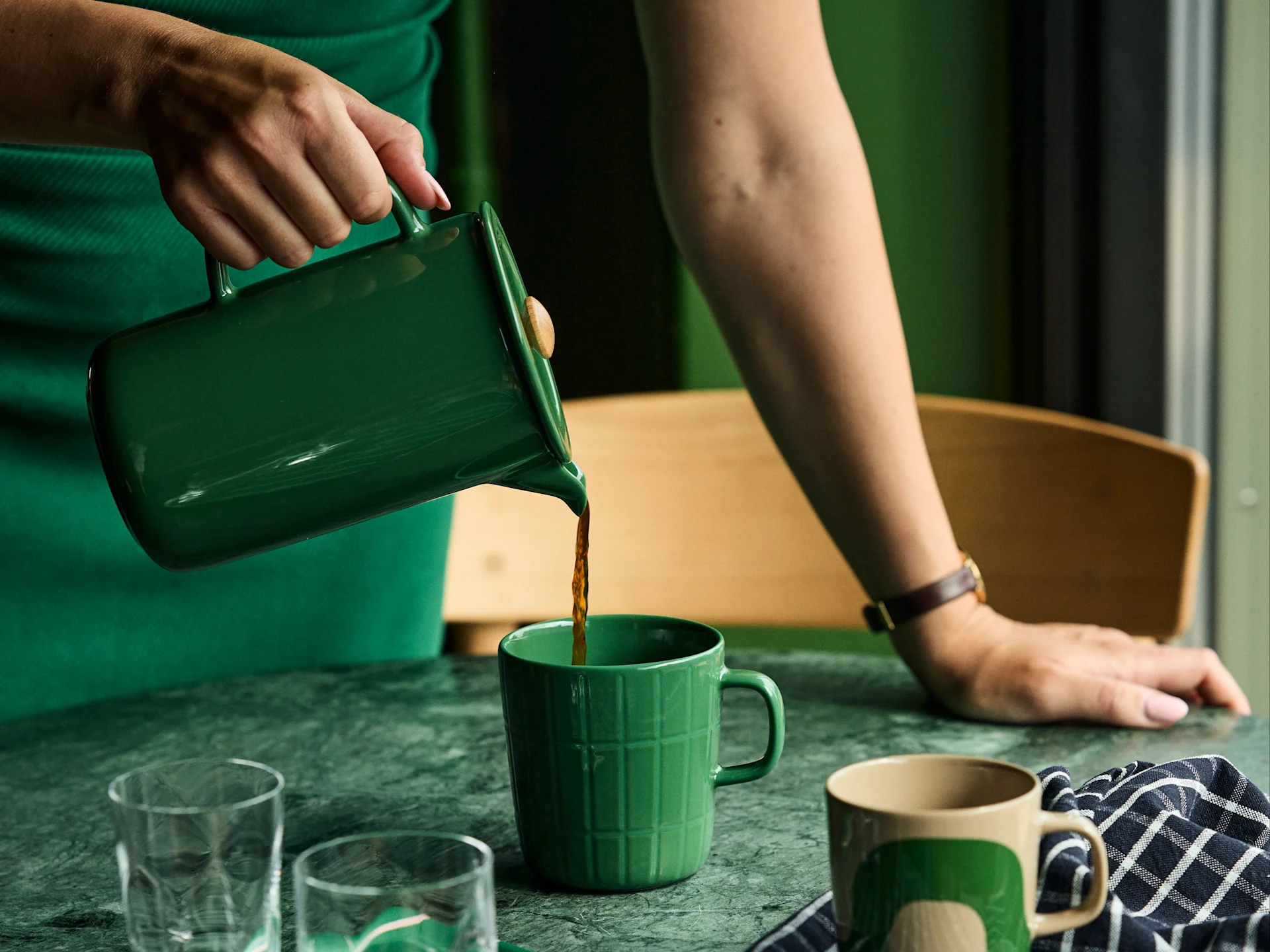 A person in a green dress pours coffee from a green pitcher into a matching green mug on a green marble table.