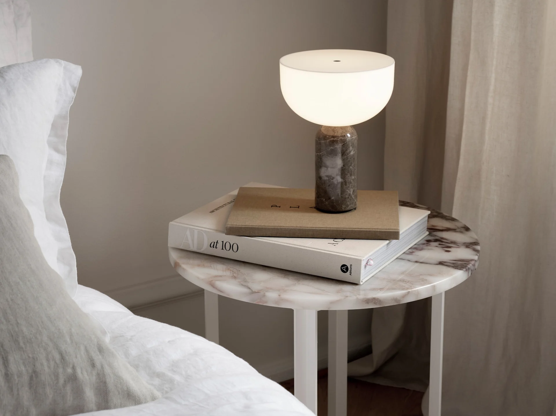 A lit table lamp with a white bowl-shaped shade and a grey marble base sits on a round marble side table with books next to a bed.
