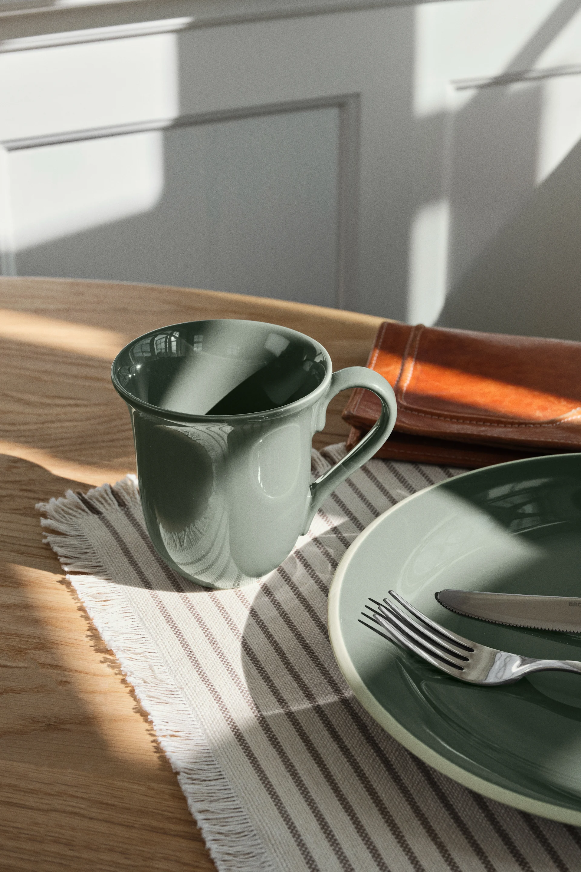 Light green ceramic mug and plate with fork and knife on a striped placemat, on a wooden table with sunlight.