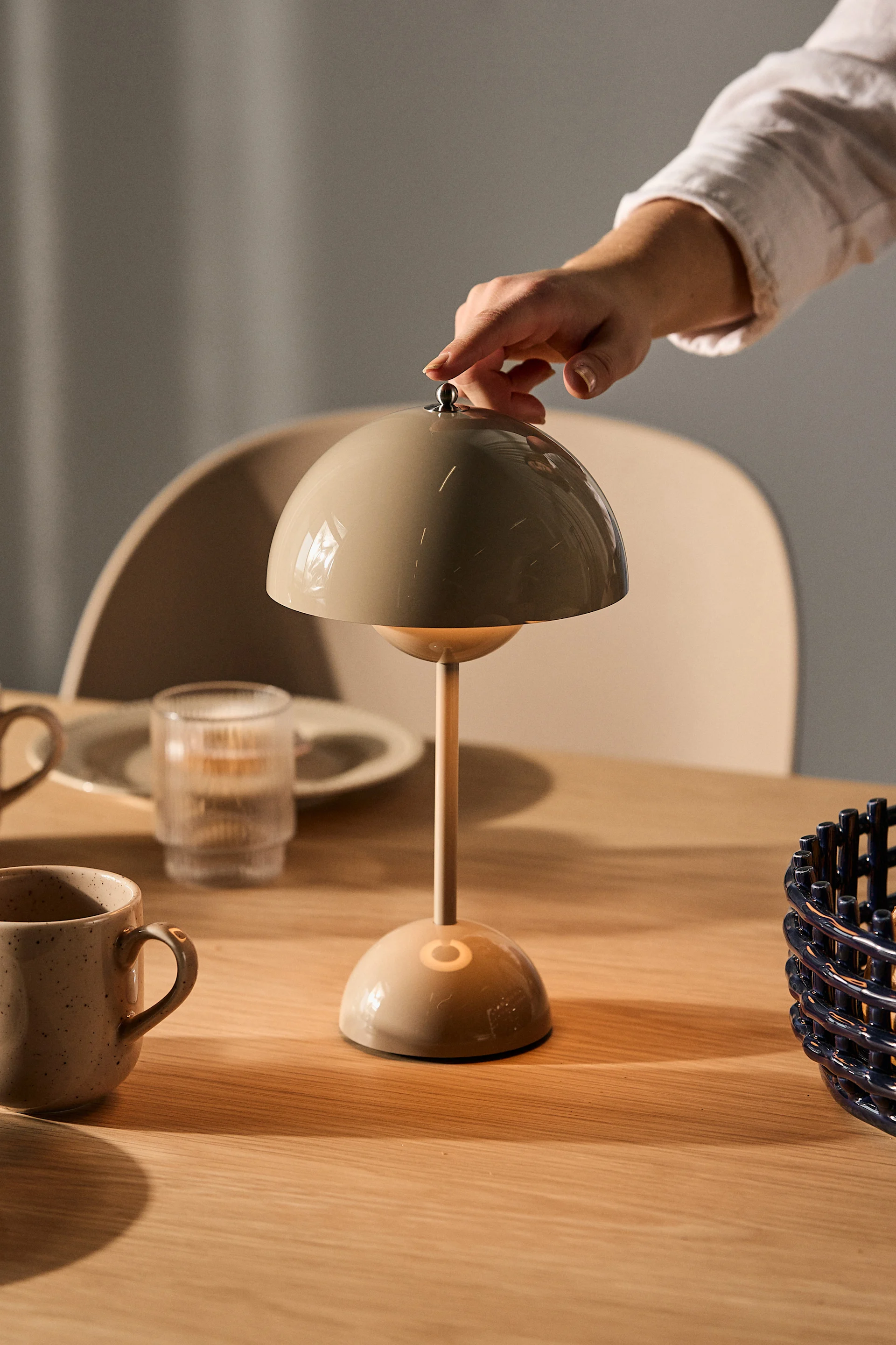 A hand adjusts a beige mushroom-shaped table lamp on a wooden table with mugs and a glass.