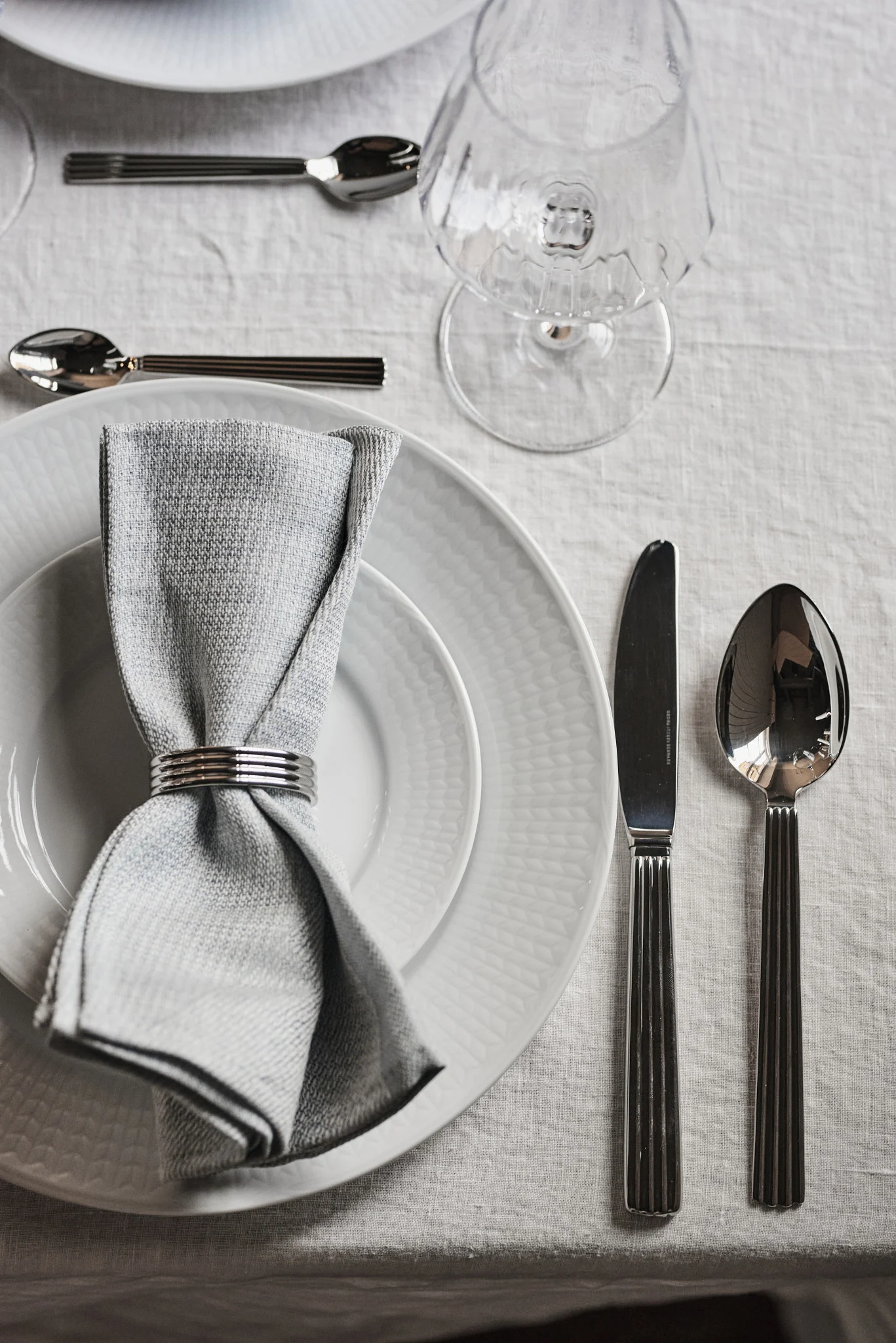 Elegant table setting with white plates, a grey napkin with a silver ring, and silver cutlery on a linen tablecloth.