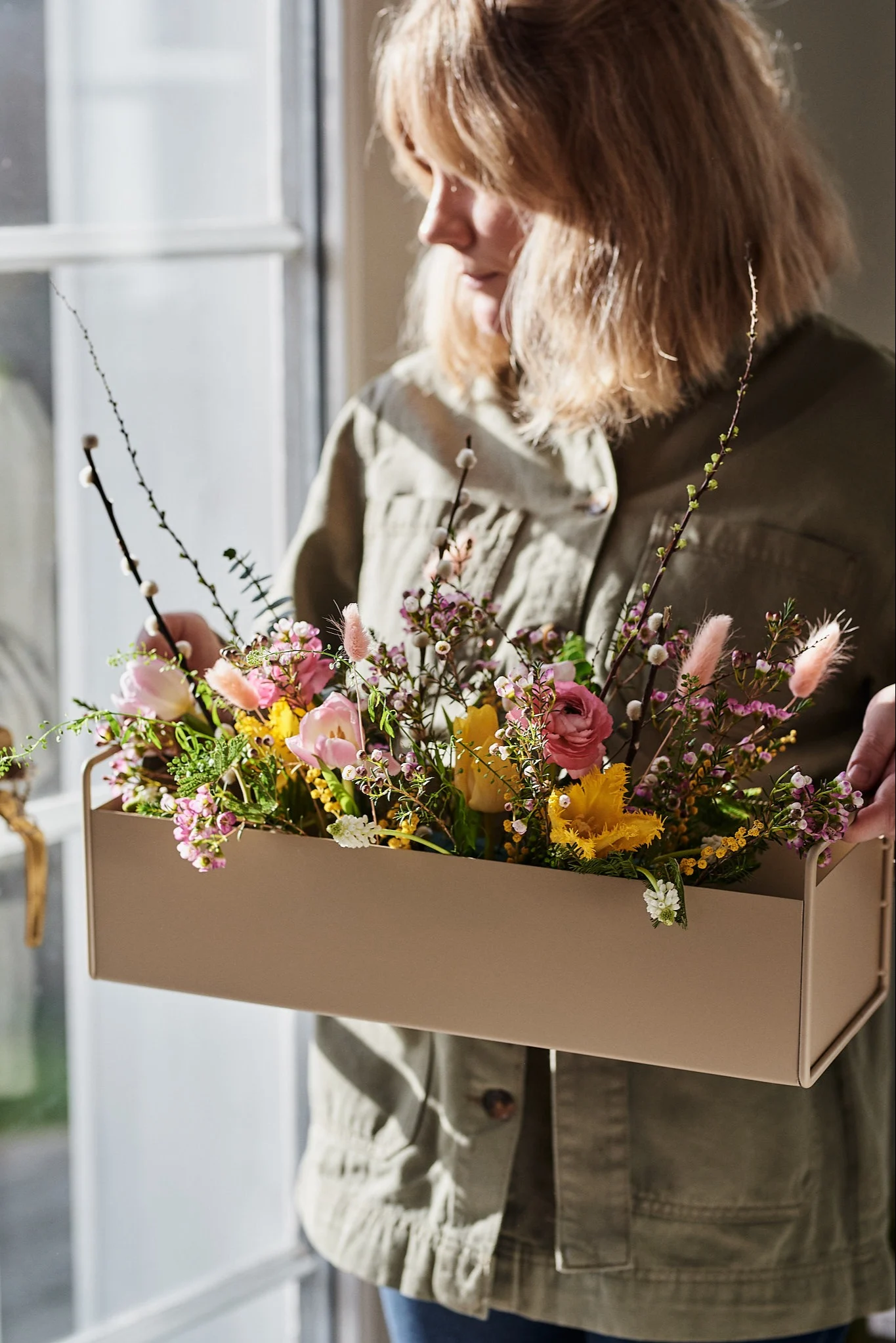 Person holding a beige rectangular planter filled with colorful spring flowers and branches.