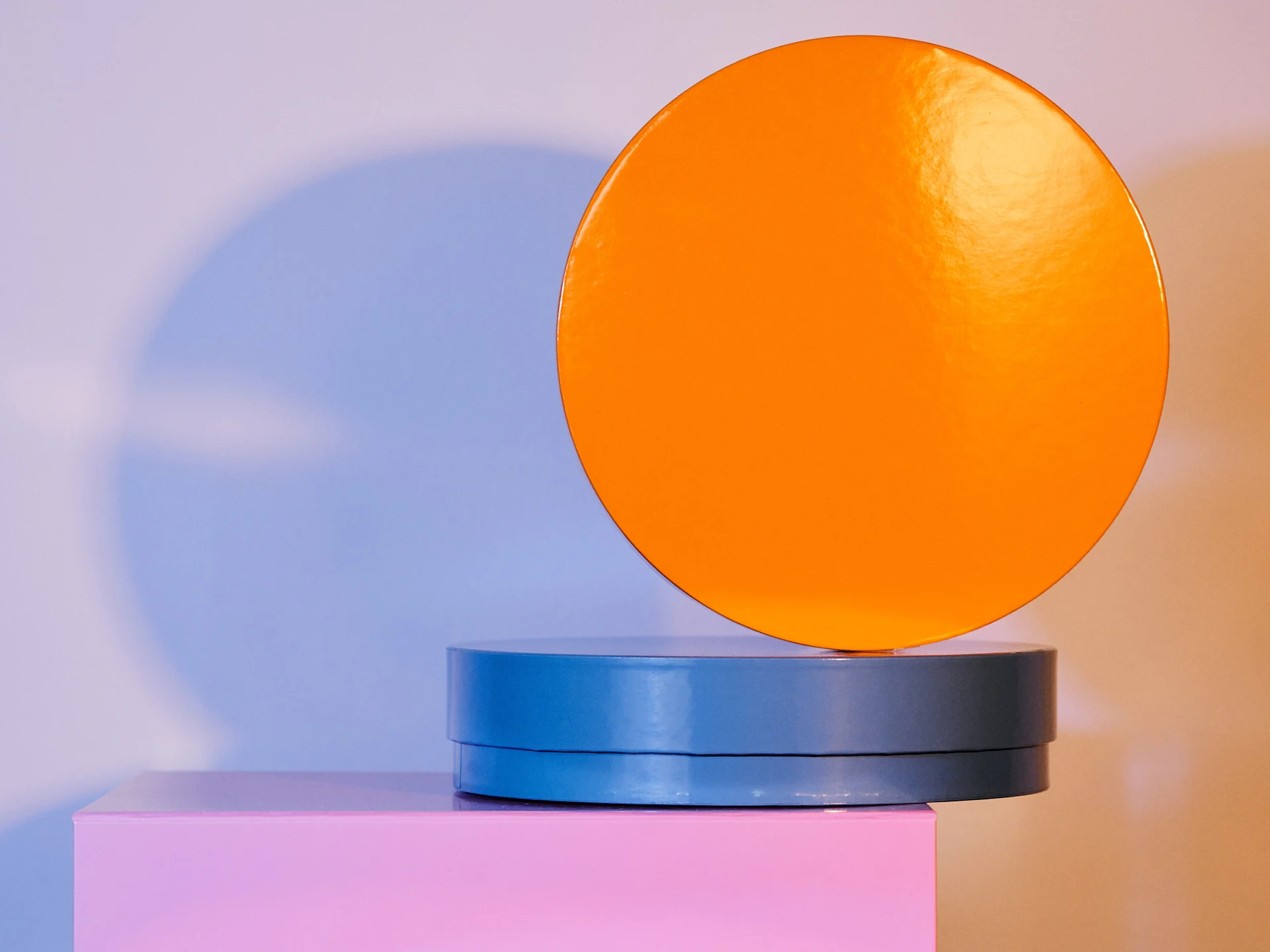 Minimalist composition with an orange circle on a blue cylinder atop a pink block.