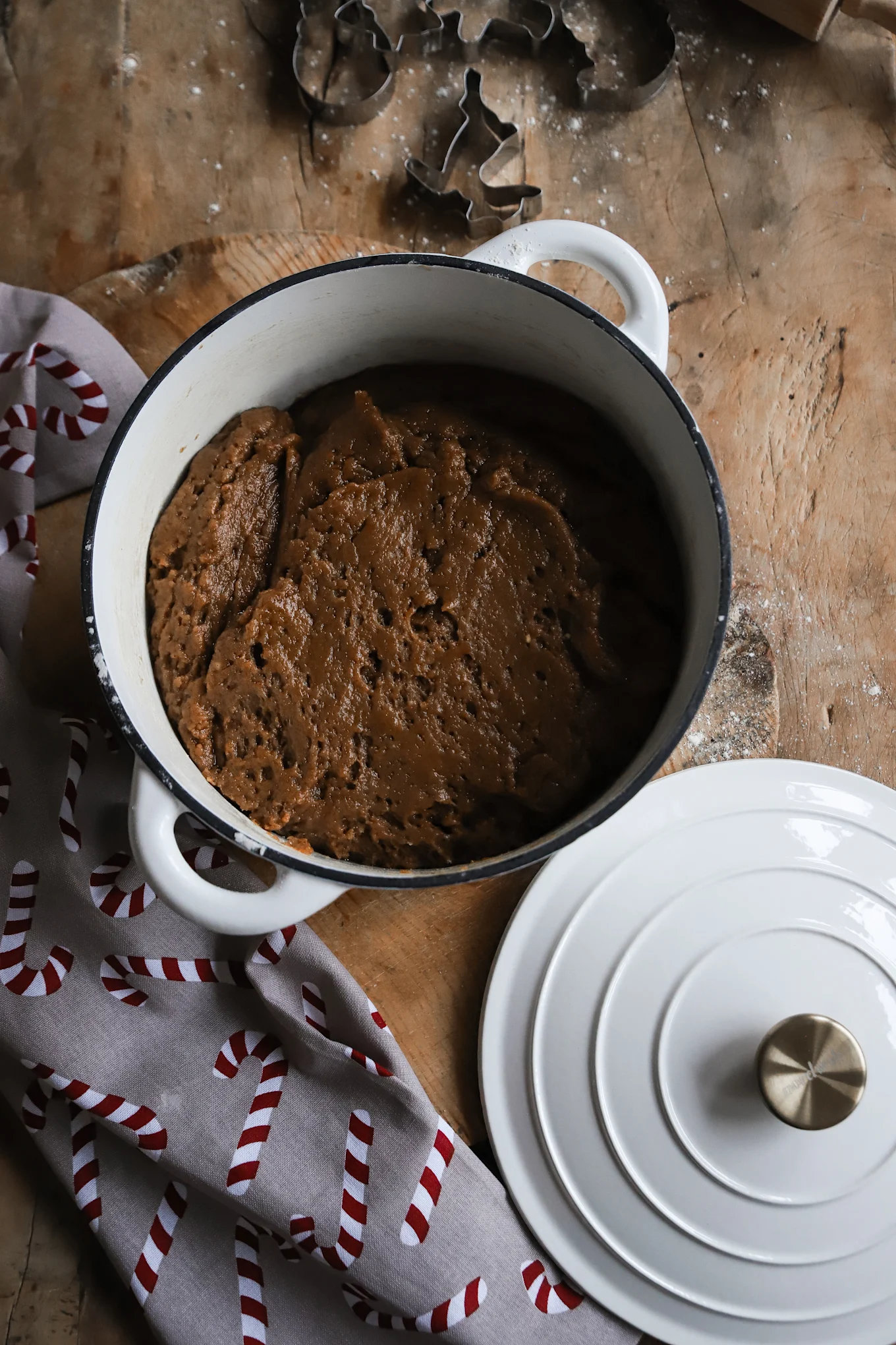 Brown cookie dough in a white pot with a lid, next to candy cane patterned fabric and cookie cutters on a wooden table.