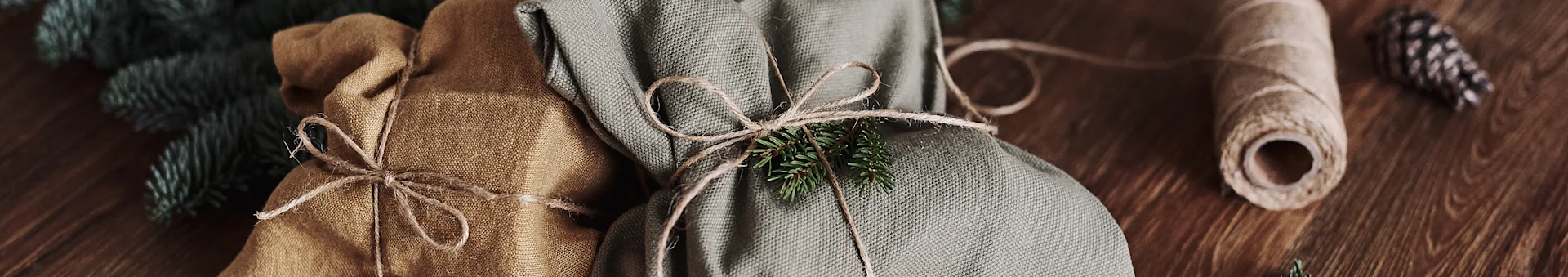 Two fabric-wrapped gifts, one brown and one green, tied with twine and fir sprigs, on a wooden table.