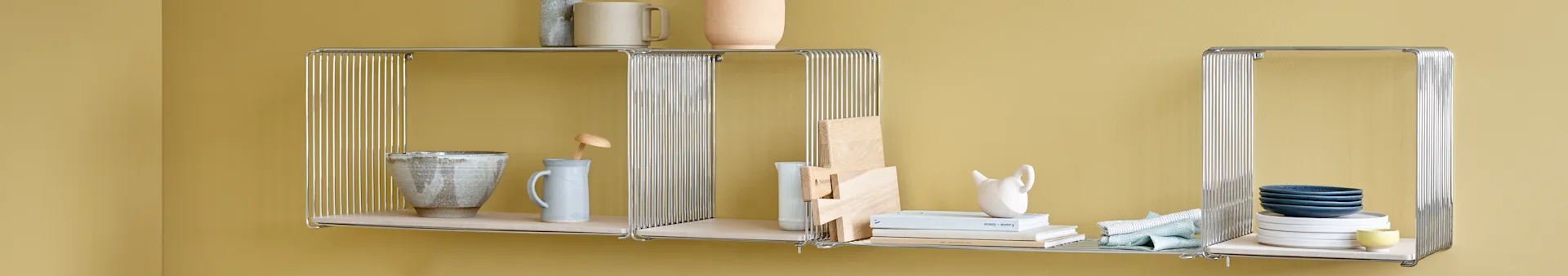 Three chrome wire and light wood wall shelves on a mustard yellow wall, displaying ceramics, cutting boards, books, and towels.