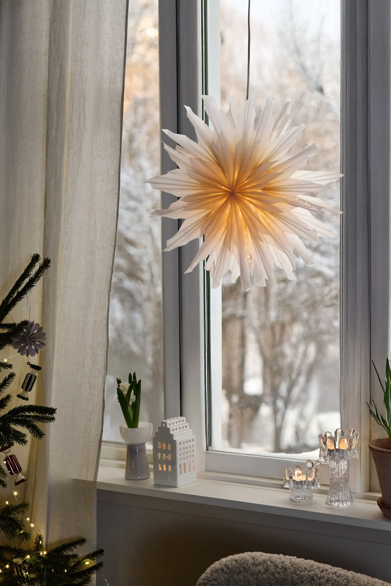 A lit white paper star hangs in a window. The sill features a flower, a ceramic house lantern, and glass candle holders.