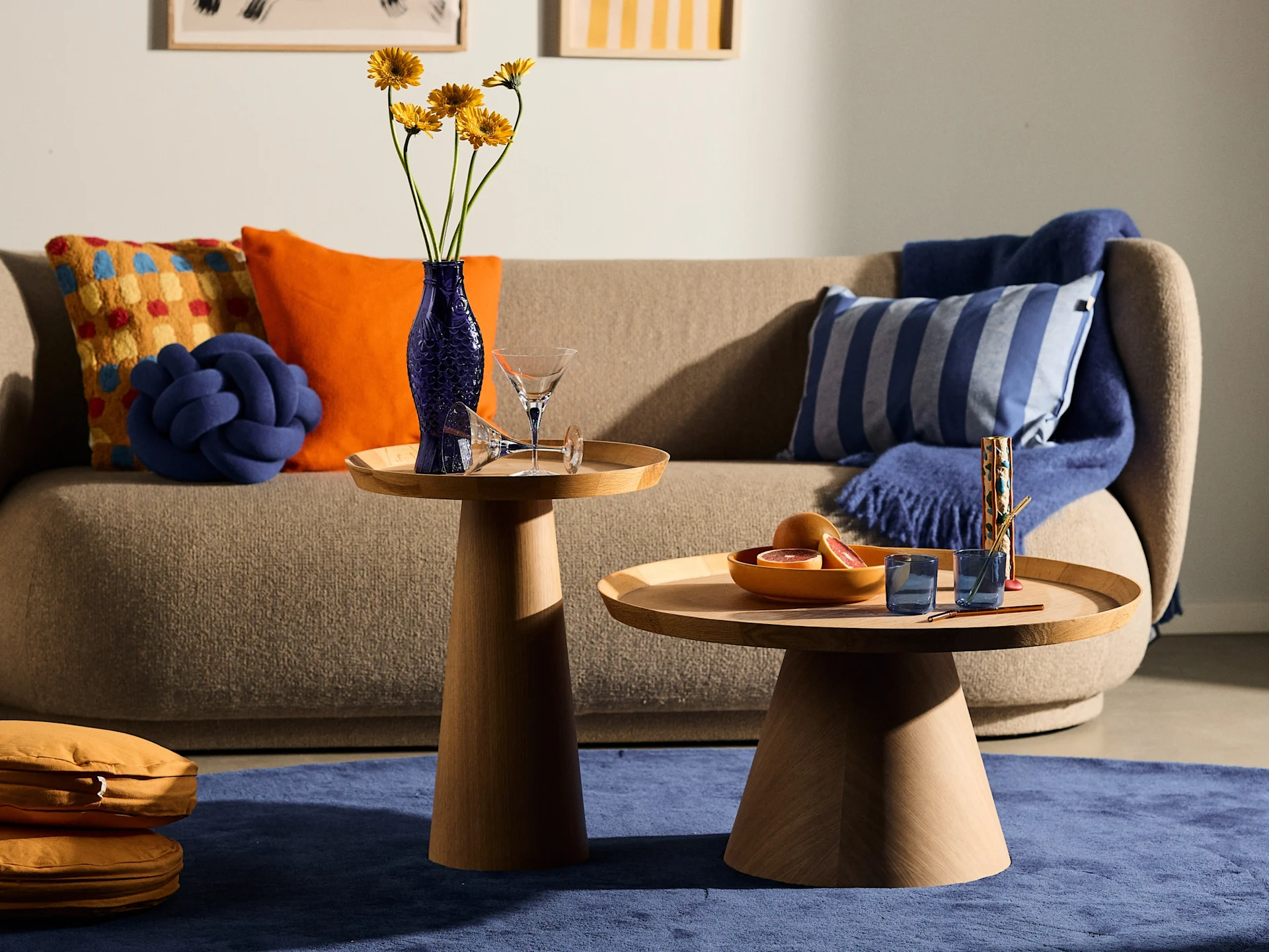 A cozy living room with a beige sofa, two round wooden side tables, a blue rug, and colorful cushions. Yellow flowers in a blue vase and grapefruit on tables.