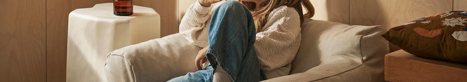 Person relaxing on a cream sofa, wearing a textured cream sweater and blue jeans, with a drink on a nearby side table.