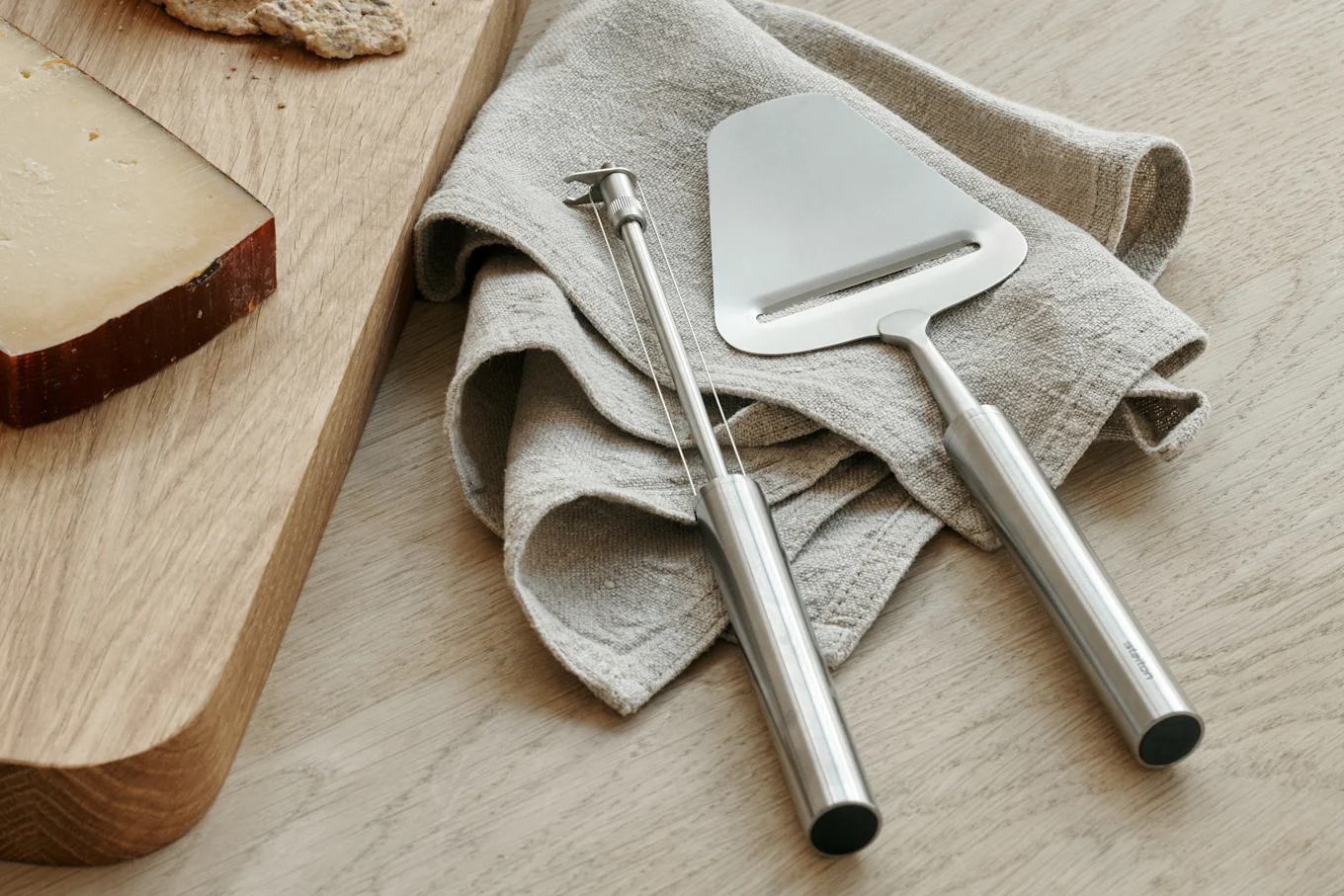 A wedge of cheese on a wooden board next to a linen napkin with a stainless steel cheese slicer and wire cutter.