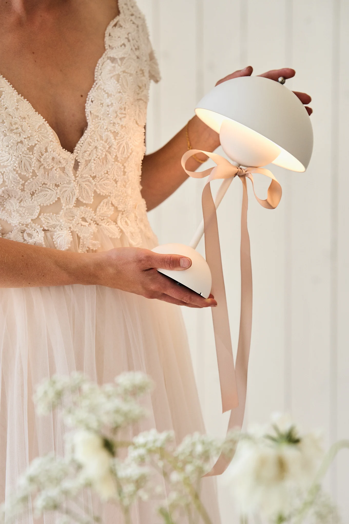 Person in a lace wedding dress holding an illuminated white modern table lamp with a beige ribbon.