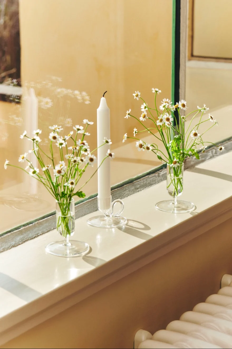 Three clear glass decorative items on a sunlit windowsill: two vases with white daisies and one tall white candle in a holder.