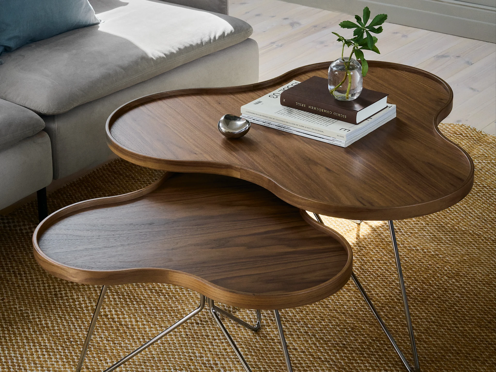 Nested dark wooden coffee tables with organic shapes and metal legs, on a textured rug. The top table holds books, a small plant, and a silver bowl.