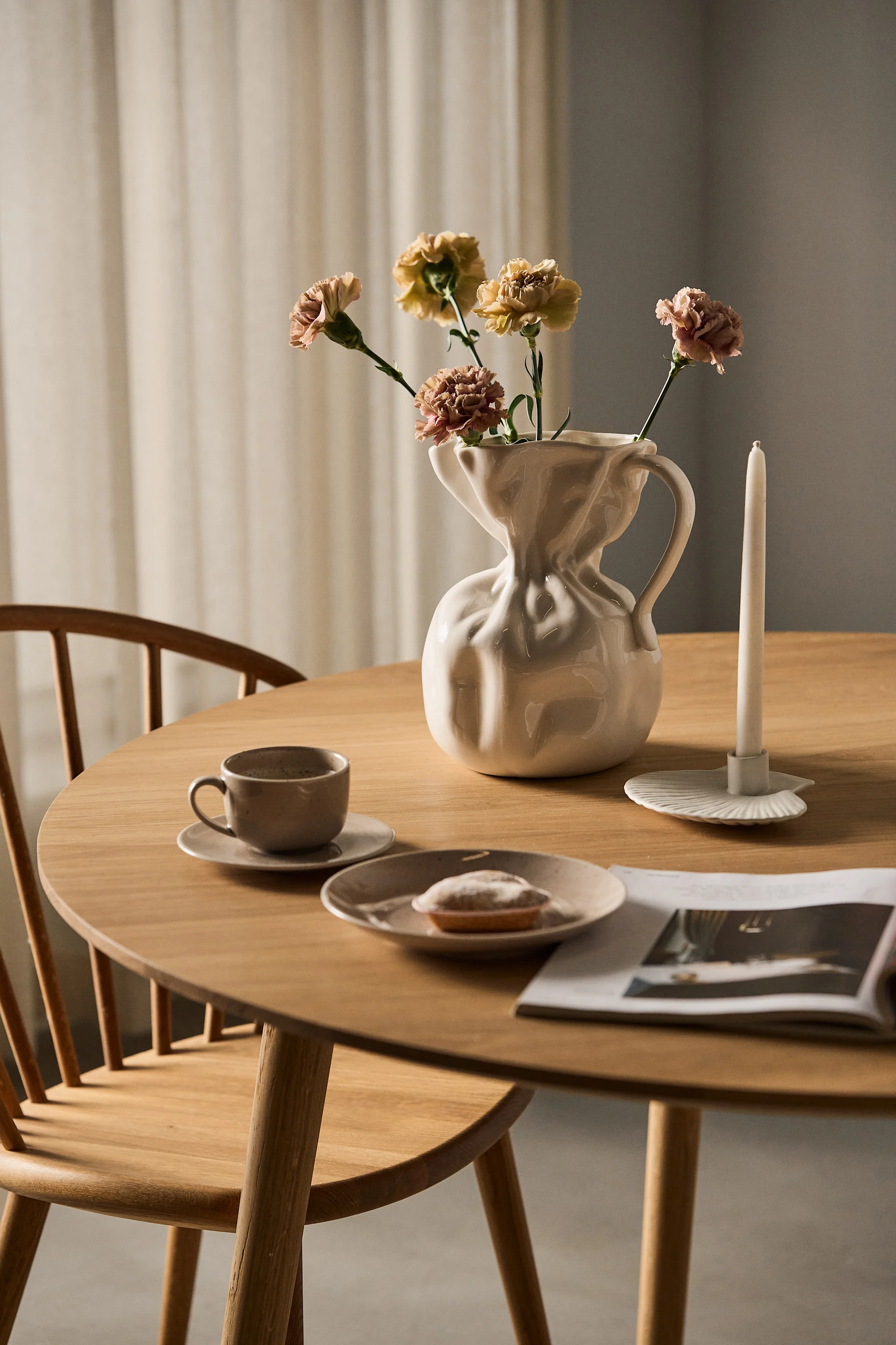 A round wooden table with a white vase of dried flowers, a white candle, a coffee cup, a pastry, and an open magazine.