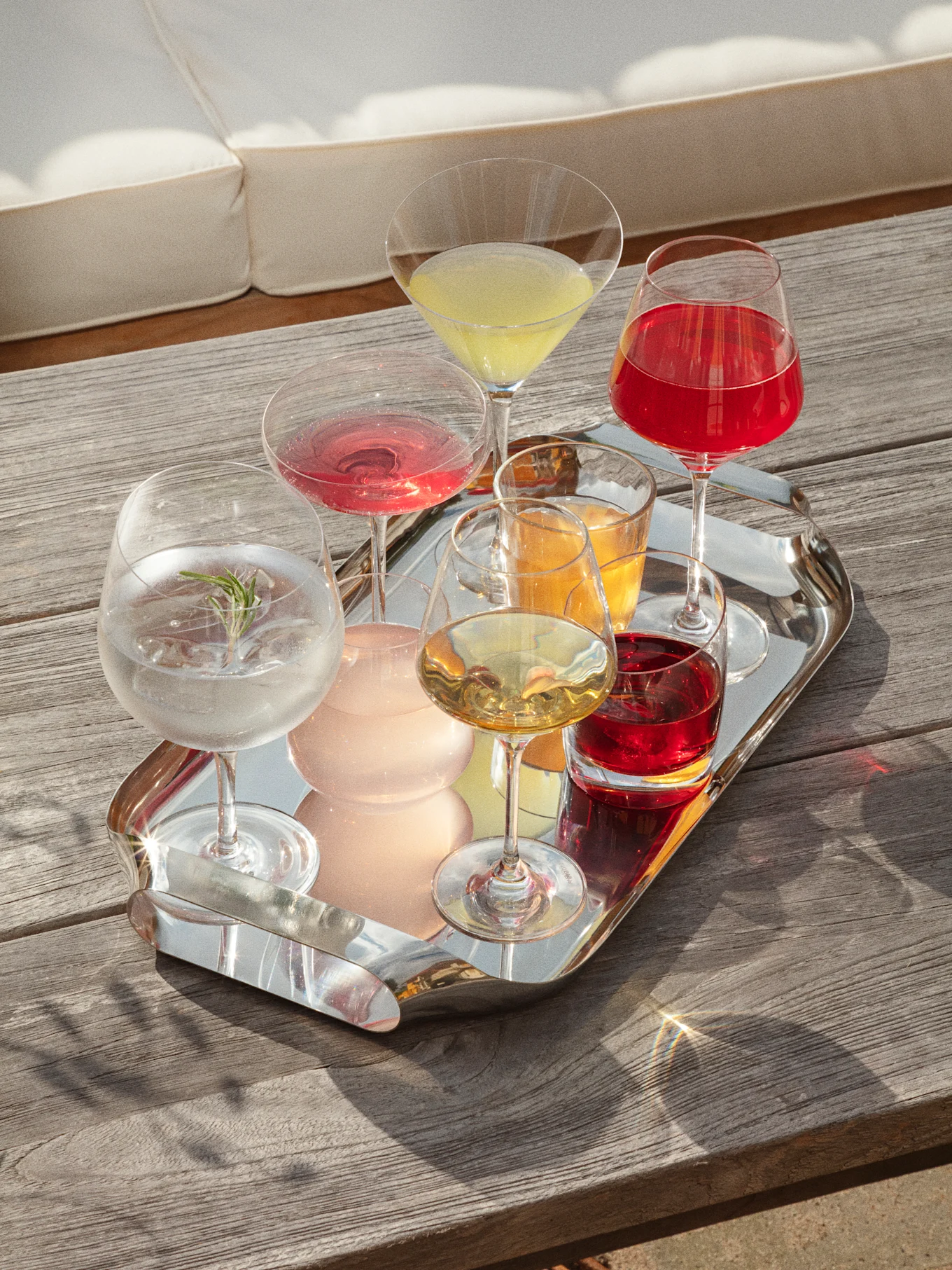 A silver tray with various colorful drinks on a wooden table, with a white sofa in the background, bathed in sunlight.