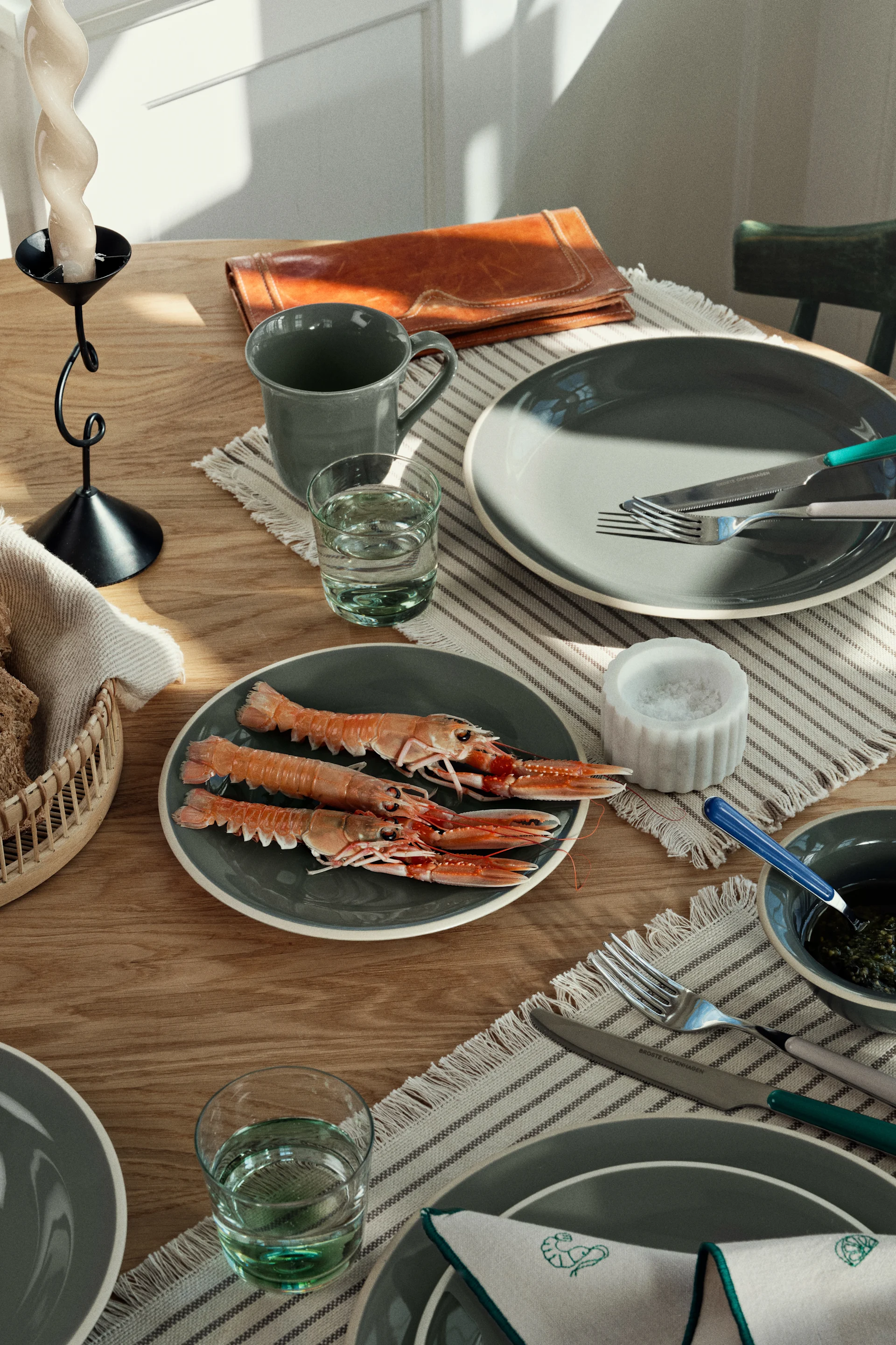 Dining table set with grey dishes, cutlery, glasses, bread, and cooked langoustines in natural light.