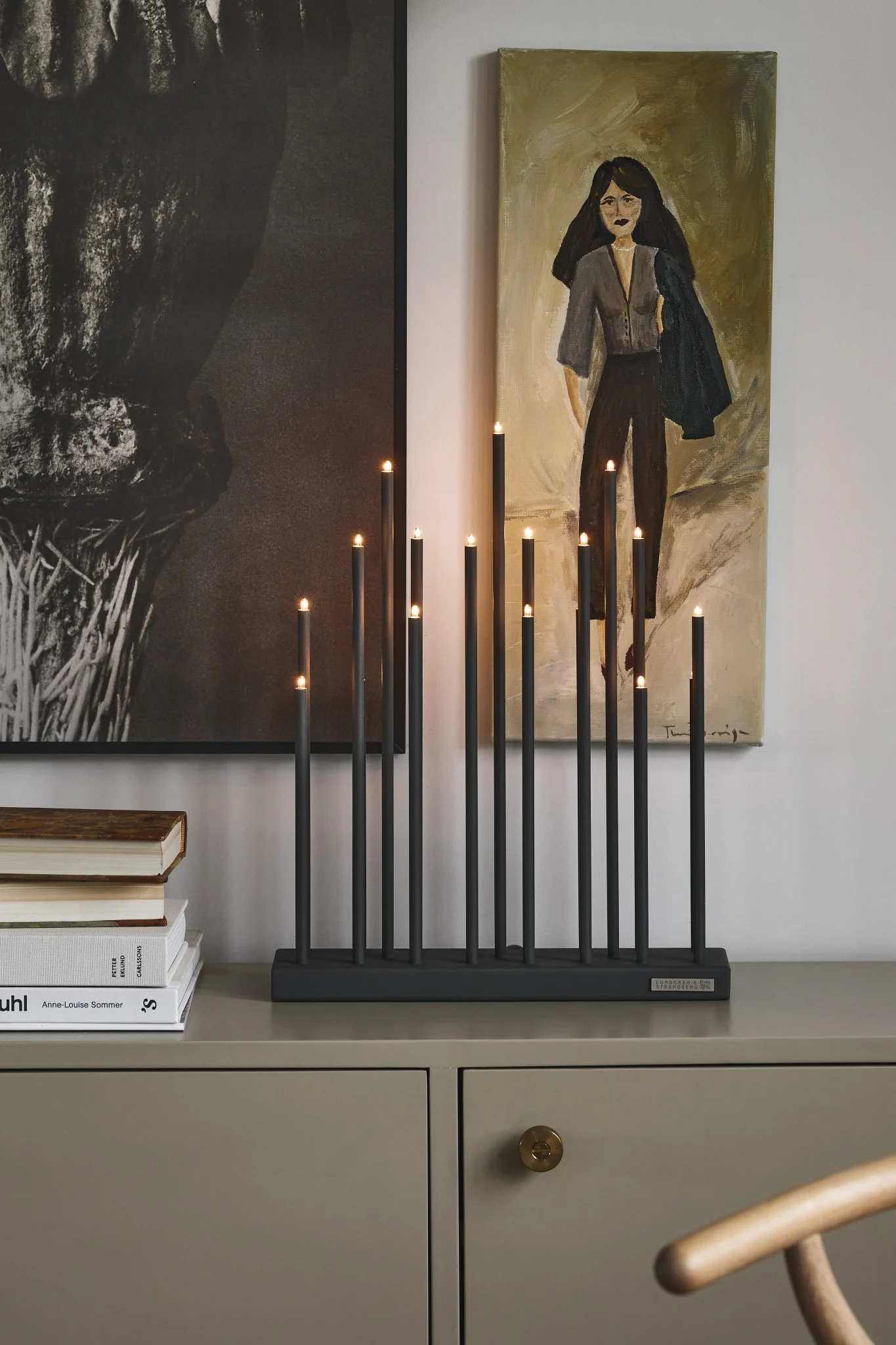Dark gray candlestick with multiple black lit candles of varying heights on a beige cabinet with artworks and books.