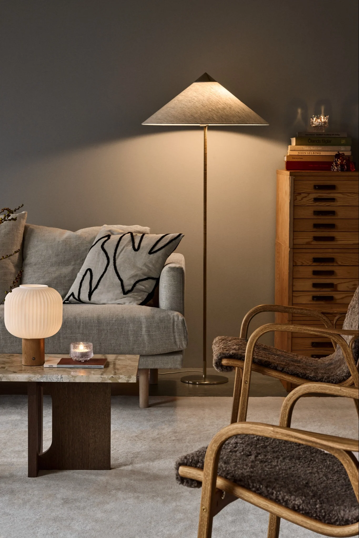 Cozy living room with a light grey sofa, ribbed table lamp, conical floor lamp, wooden chairs with fur, and a cabinet with books.