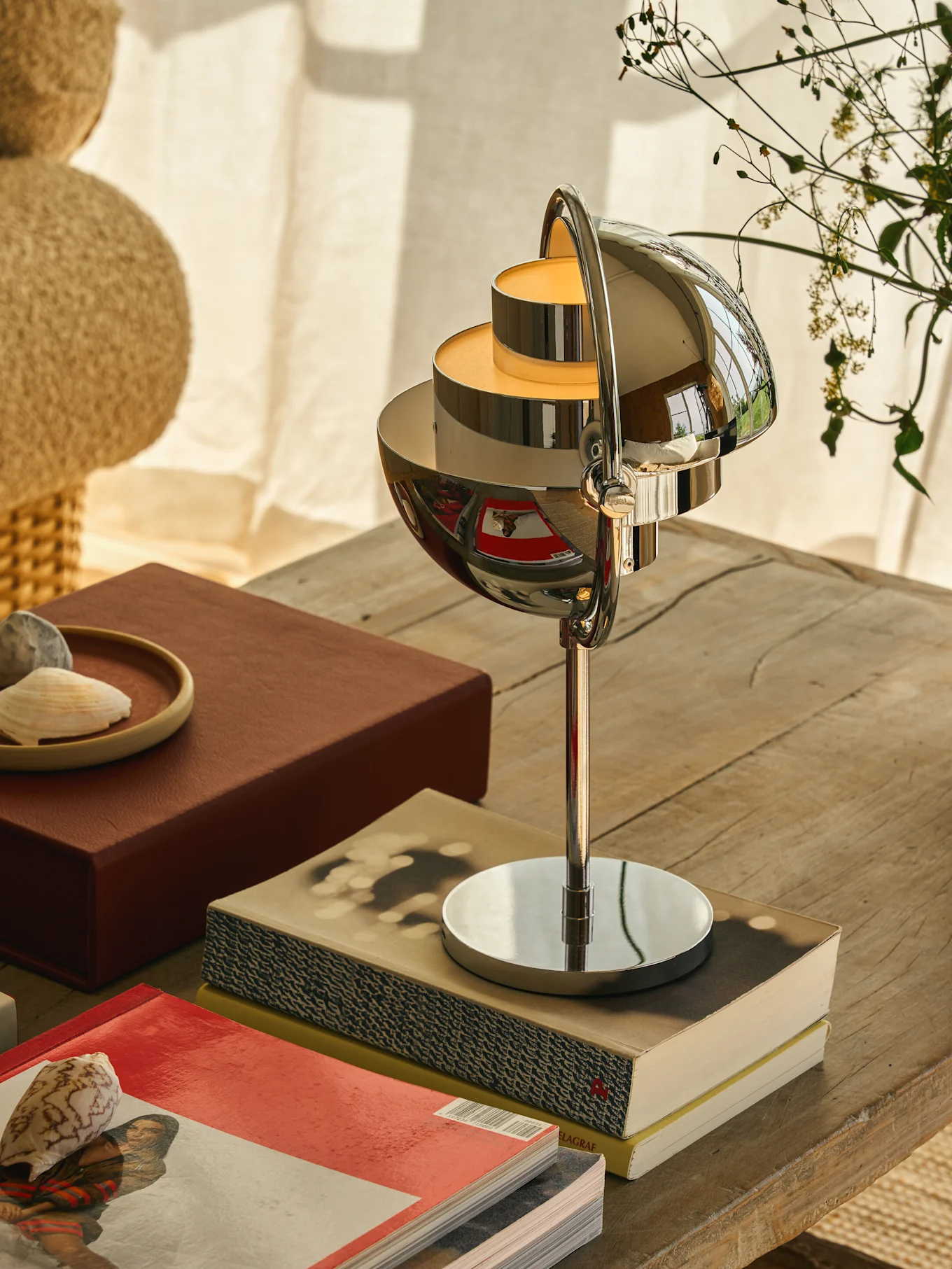 Chrome-plated table lamp with multi-tiered, spherical shades casting a warm glow, on a wooden table with books and shells.