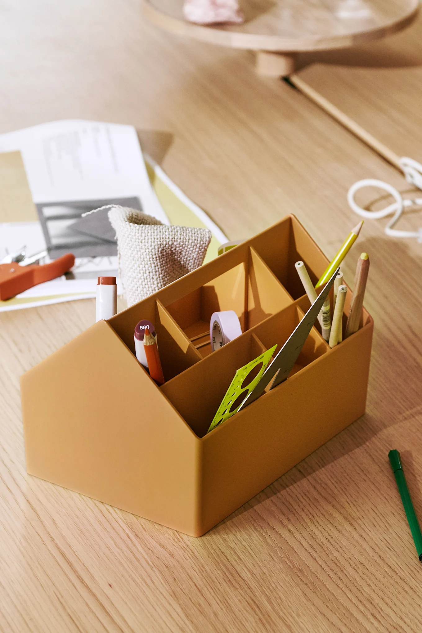 An orange desk organizer filled with pencils, a ruler, and tape on a light wood desk.