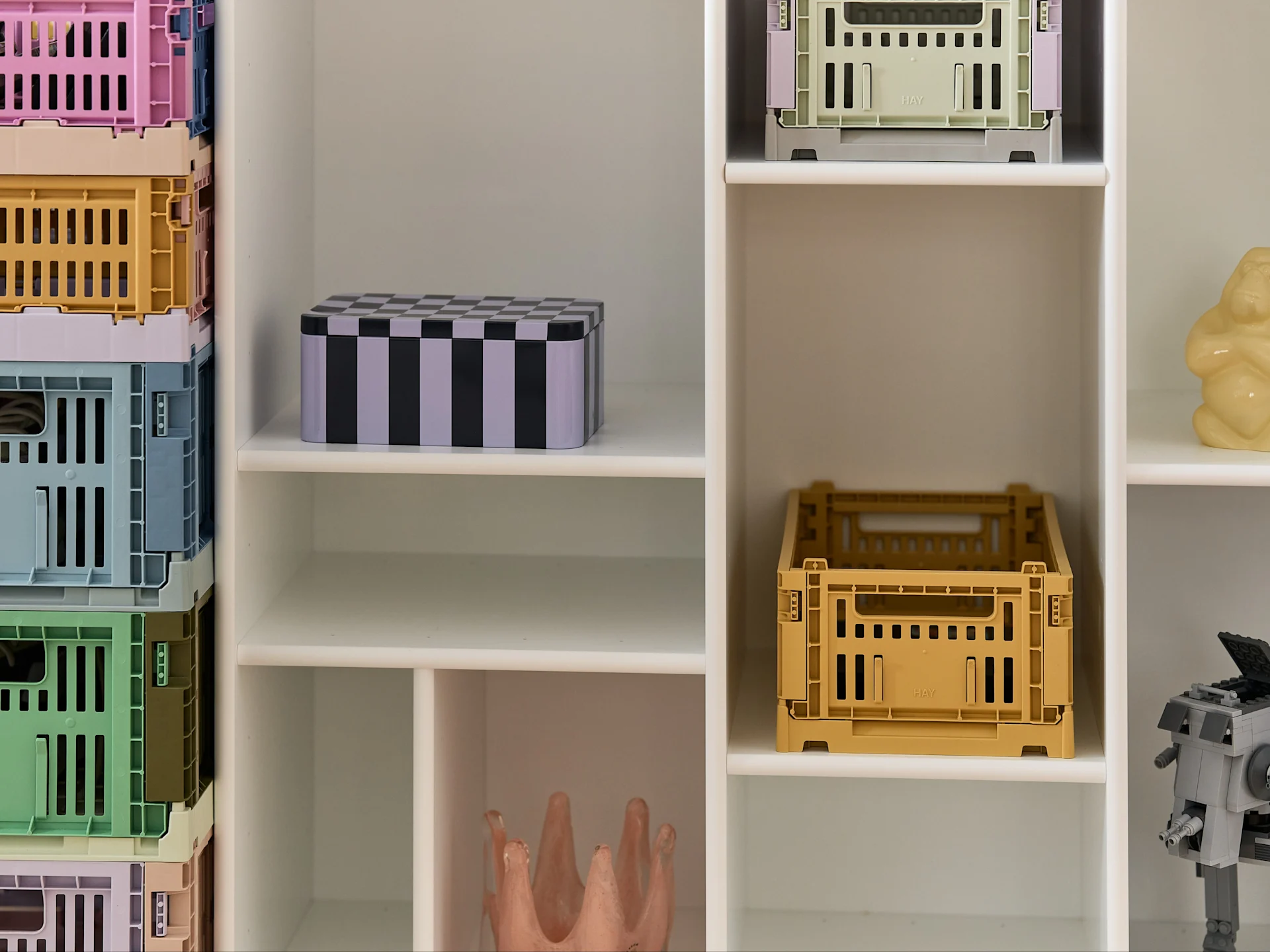 A white shelving unit filled with colorful, stacked foldable storage crates, a striped box, a yellow figure, and a gray Lego robot.