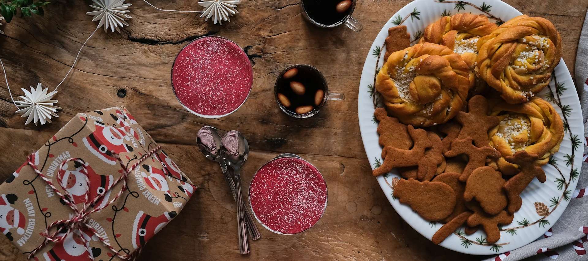 Christmas flat lay with saffron buns, gingerbread, red drinks, and a Santa gift on a wooden table.