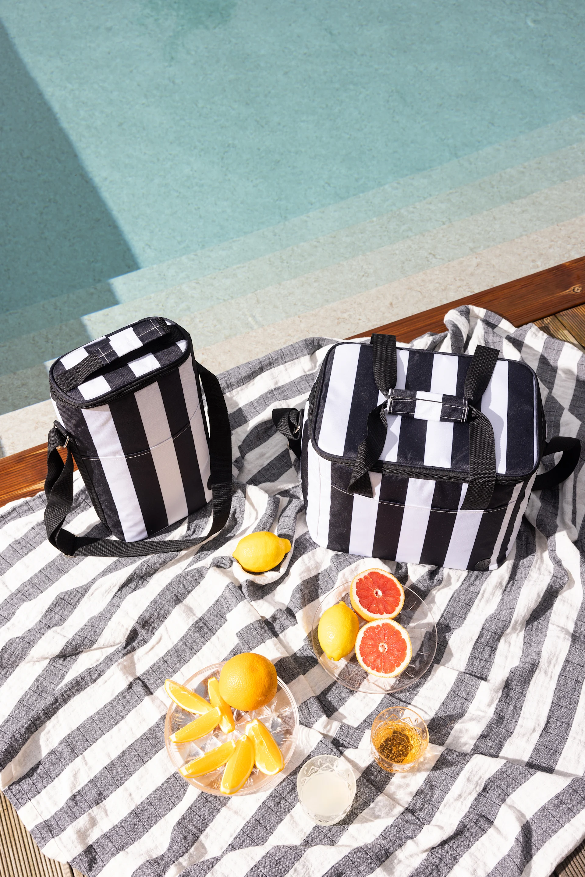 Two black and white striped cooler bags on a striped towel next to a pool, with citrus fruits and drinks.