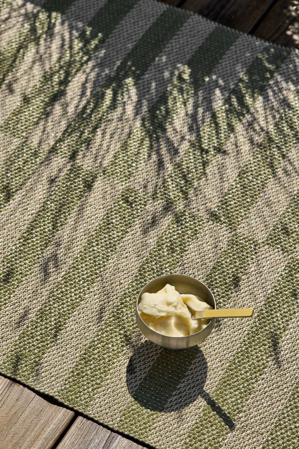 A silver bowl of pale yellow ice cream with a golden spoon sits on a green and beige striped mat with leaf shadows, next to wooden planks.