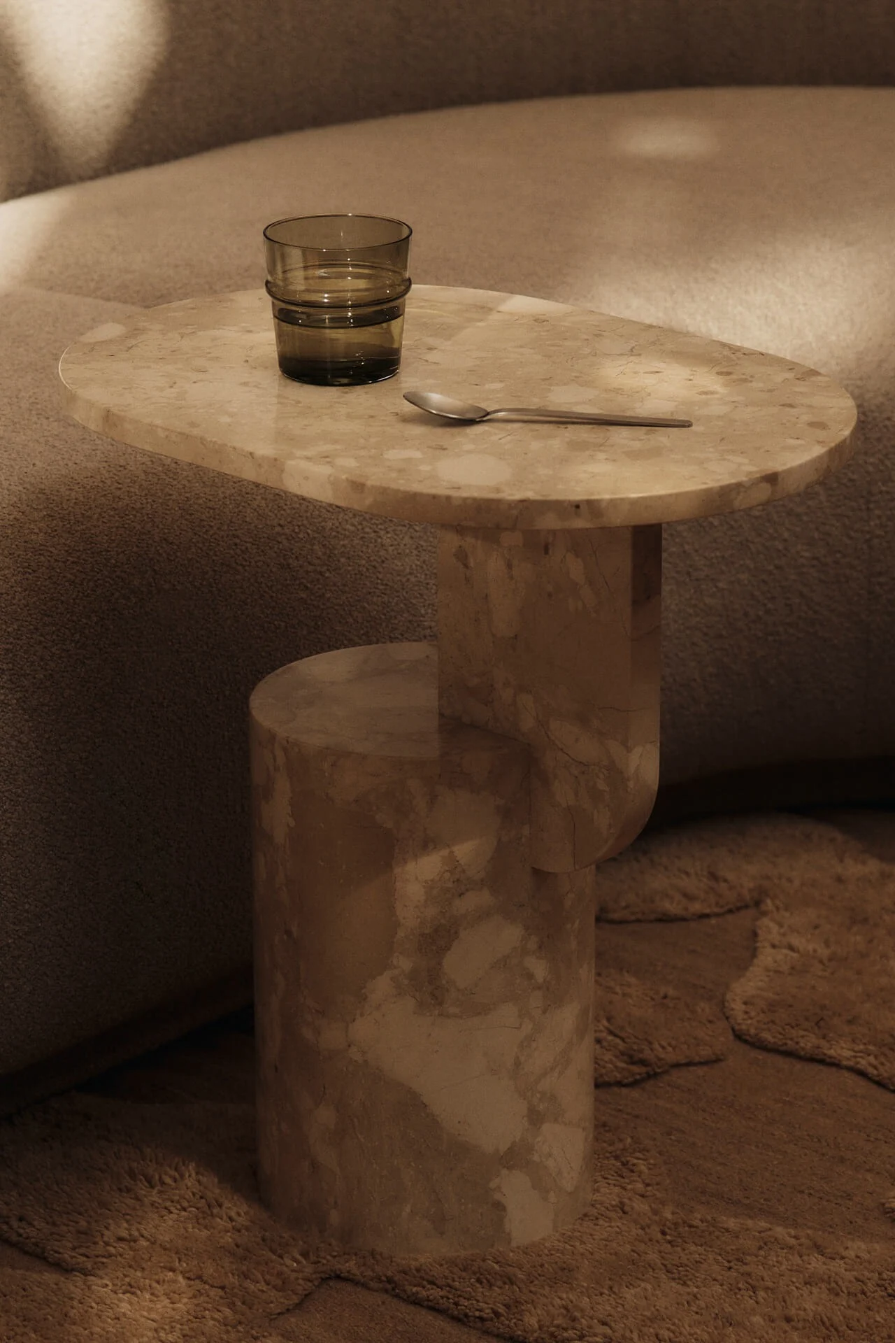 Beige marble side table with a dark green glass of water and a silver spoon, beside a textured sofa and rug.
