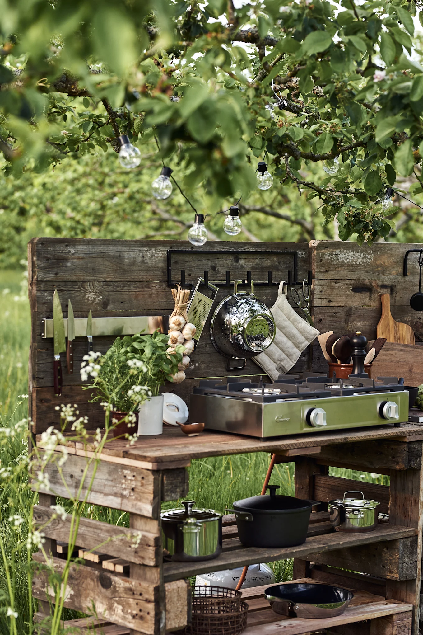 An outdoor kitchen made of rustic wooden pallets with a green portable gas stove, cooking utensils, and string lights under a leafy tree.