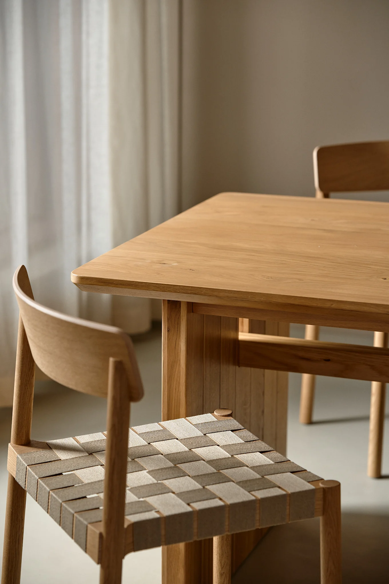 A close-up of a light wooden dining table with a chair featuring a woven seat in the foreground. Another wooden chair is blurred in the background, all against a softly lit room with sheer curtains.