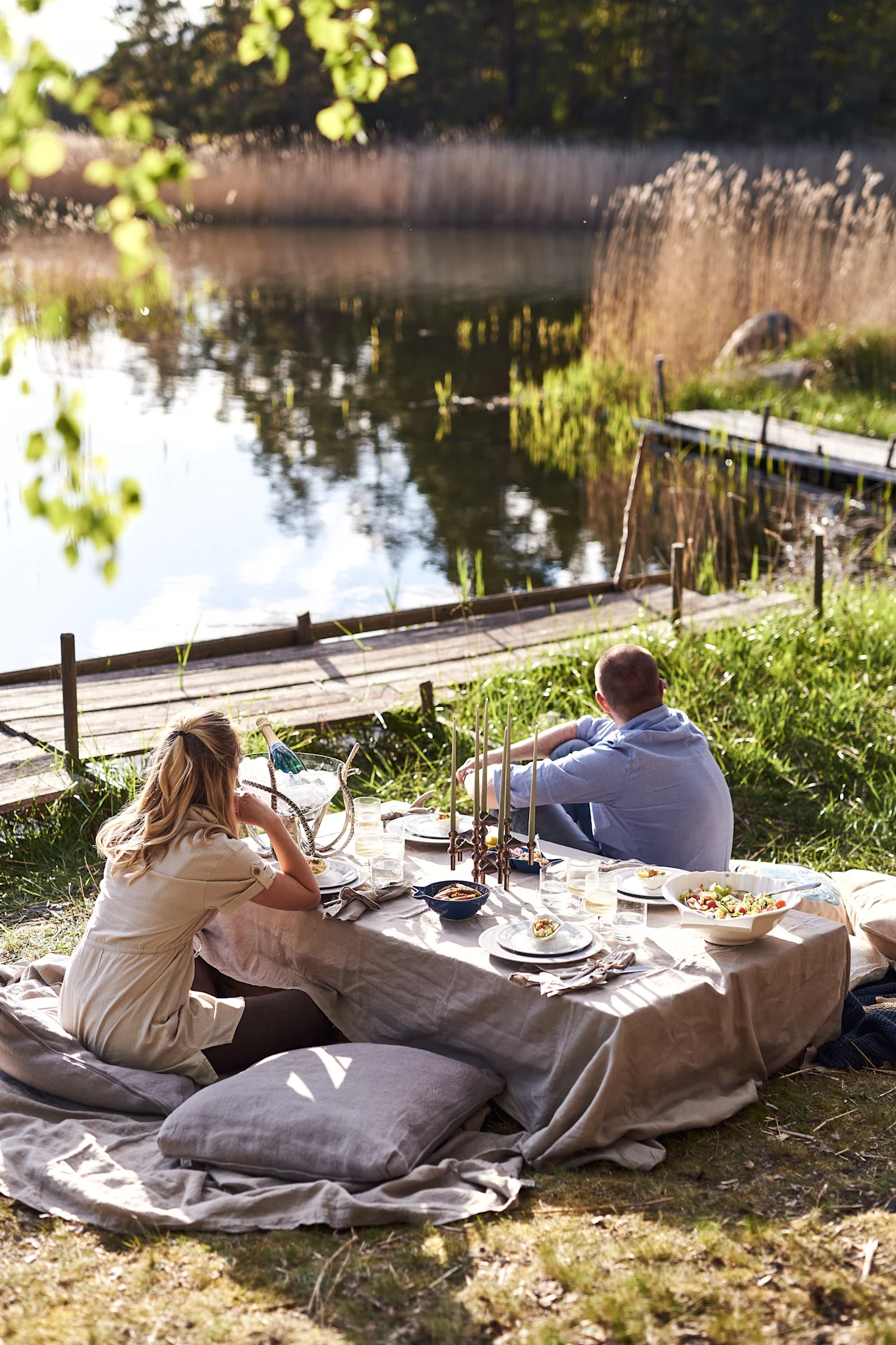 Couple enjoying a picnic by a serene lake with a low table, cushions, food, and drinks.