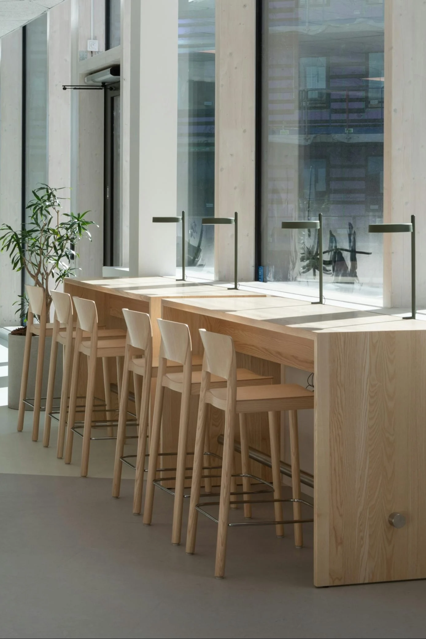 A long light wooden table with several matching bar stools and dark green desk lamps, illuminated by natural light from large windows.