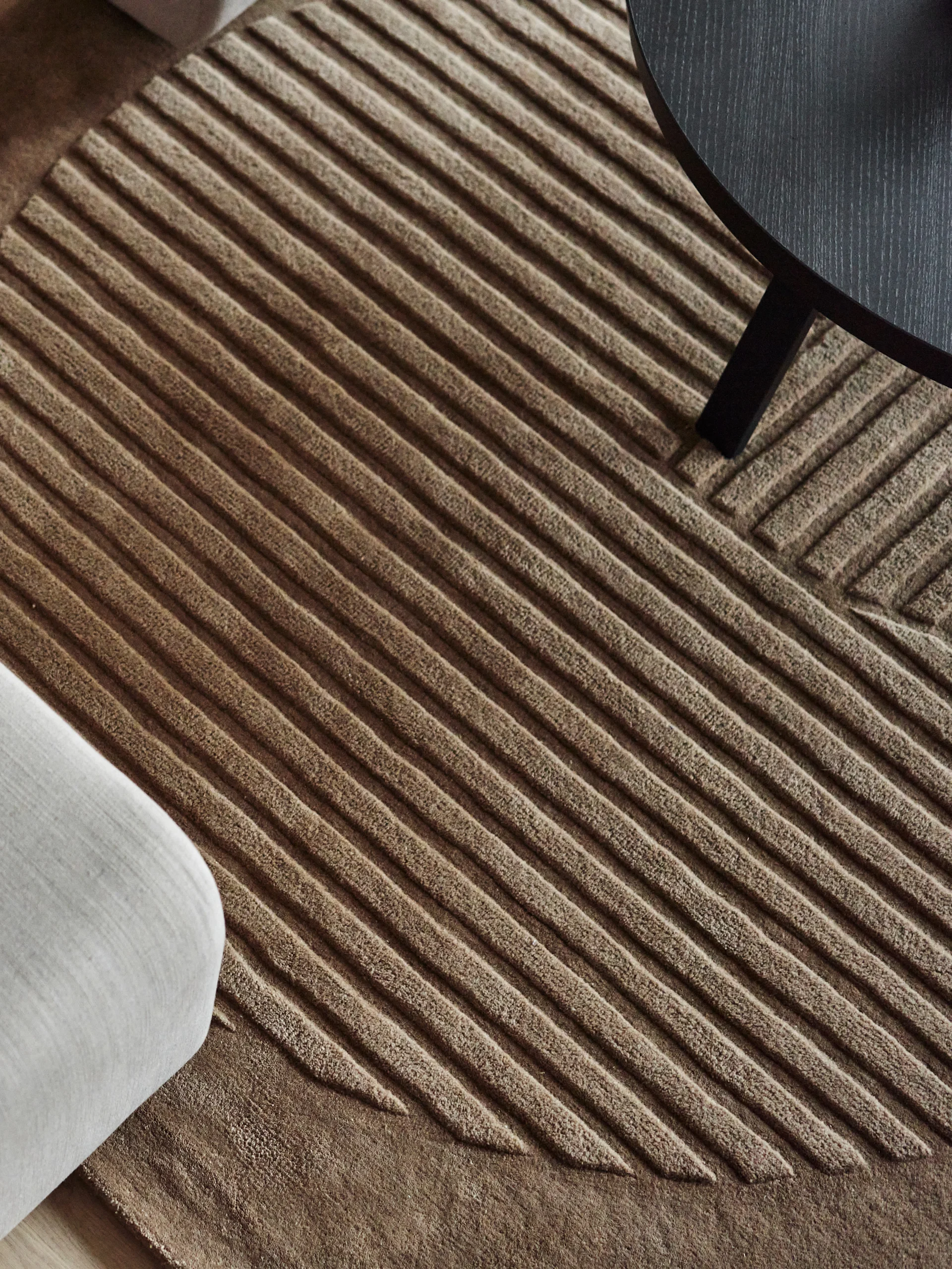 Overhead view of a light brown ribbed rug featuring diagonal lines, with a white sofa and dark round table visible.