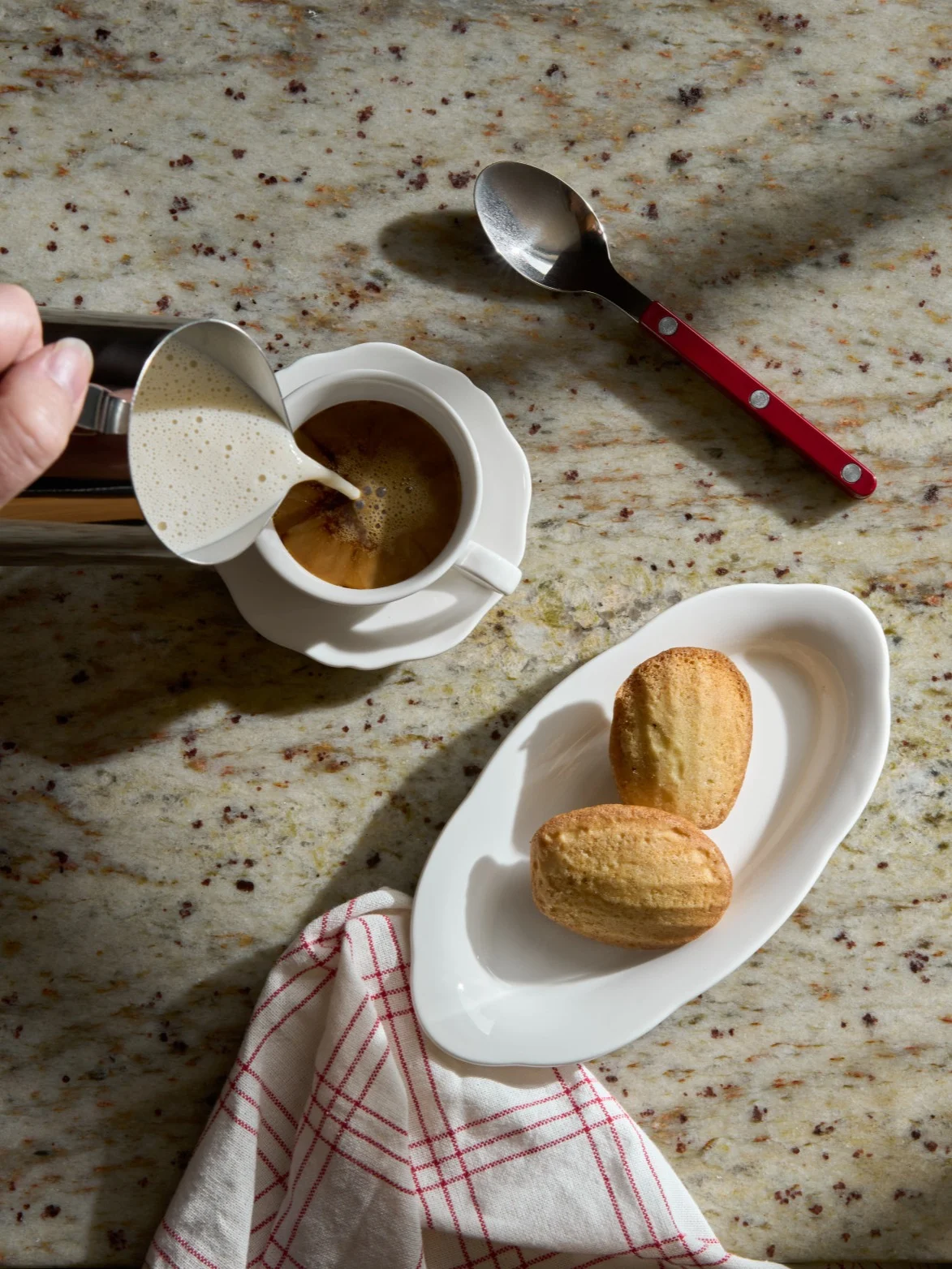 Hand pouring frothed milk into a coffee cup next to madeleine cookies on a granite counter.