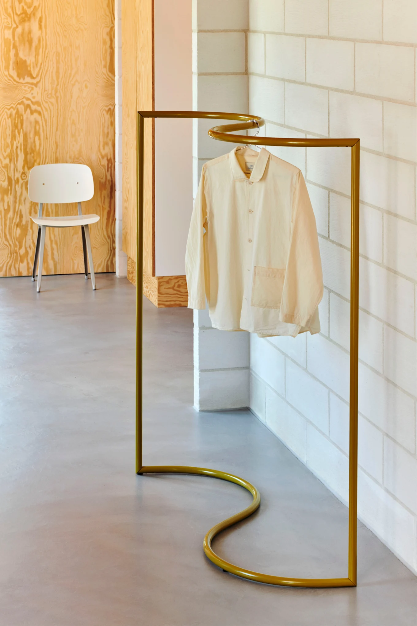 A light cream long-sleeved shirt hangs on a golden, S-shaped clothing rack against a background of wooden panels and concrete blocks.