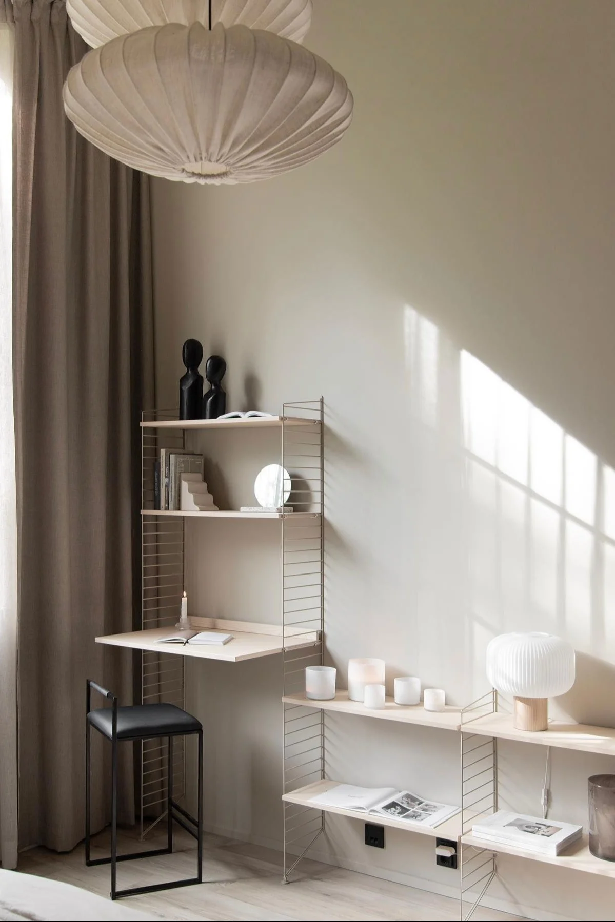 Modern home office: light wood shelving system with desk, black stool, and decor. Sunlight casts shadows.