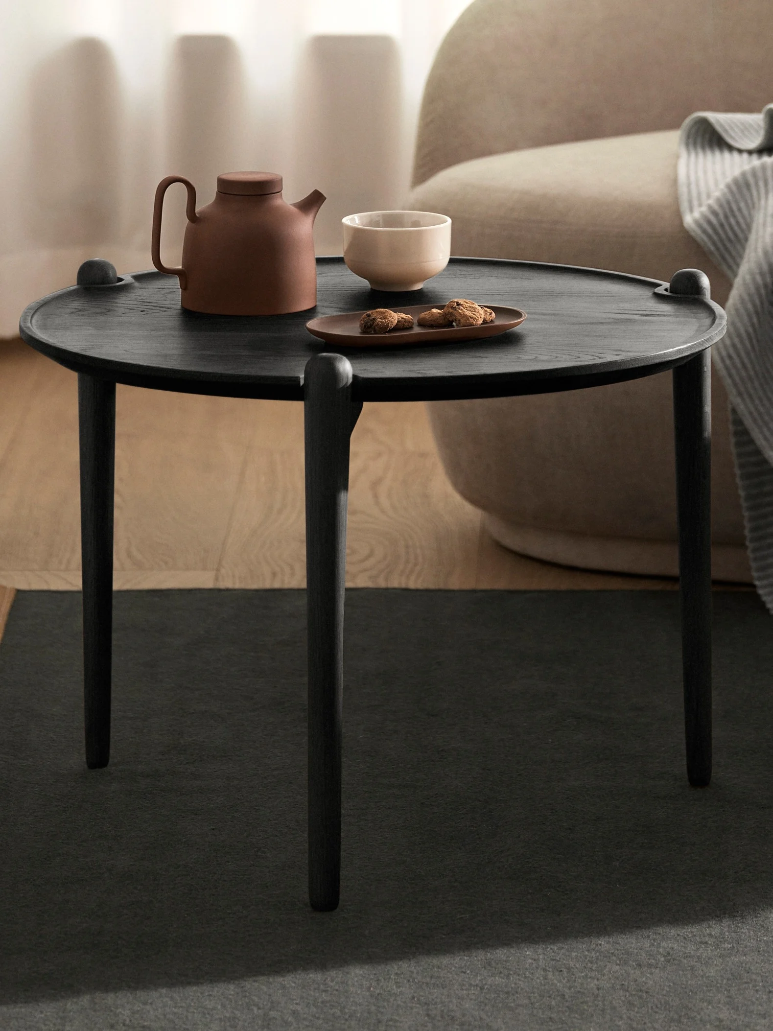 A dark round wooden side table with a brown teapot, a light bowl, and cookies on a plate, next to a beige armchair.