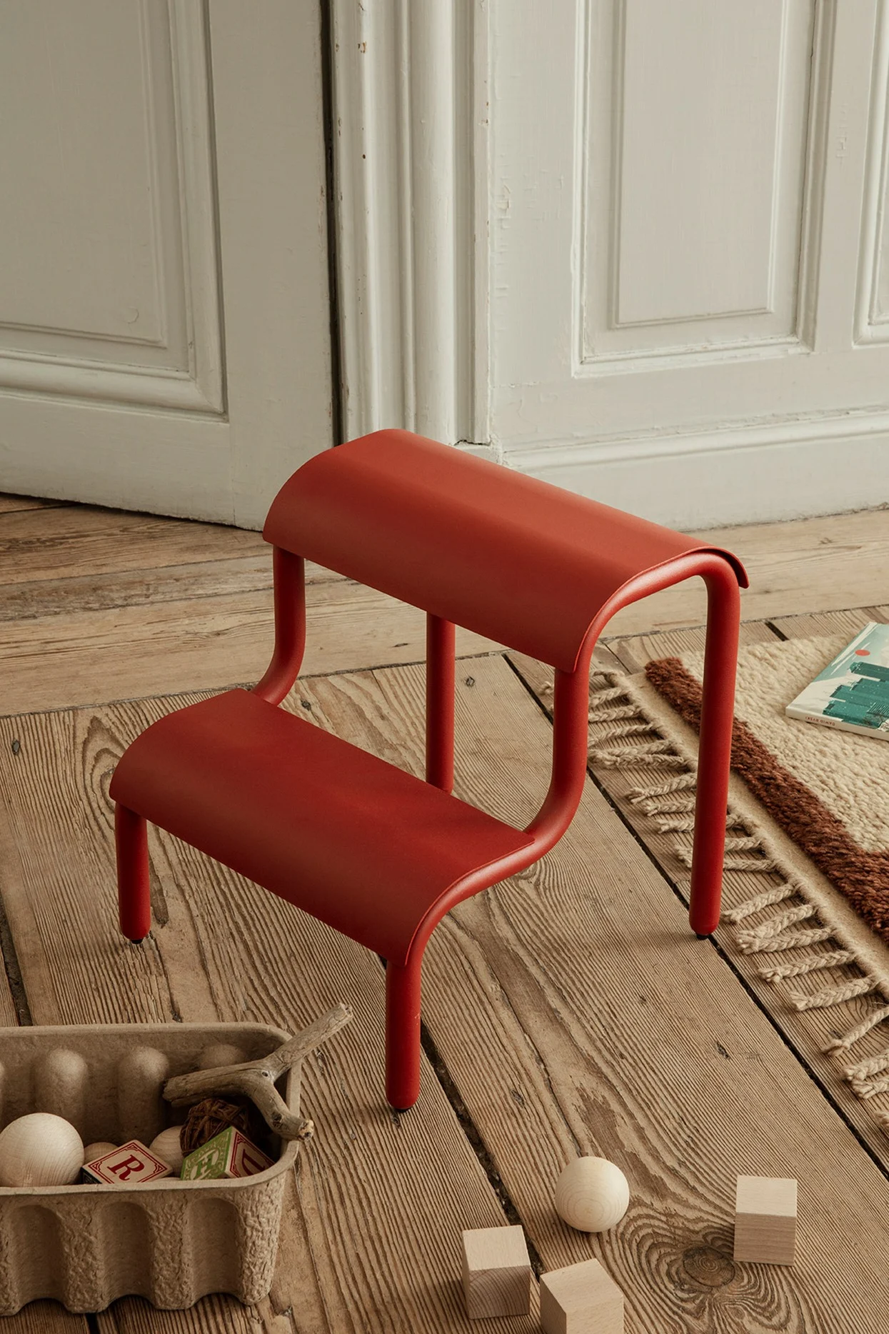 Red curved two-step stool on wooden floor, with toys.